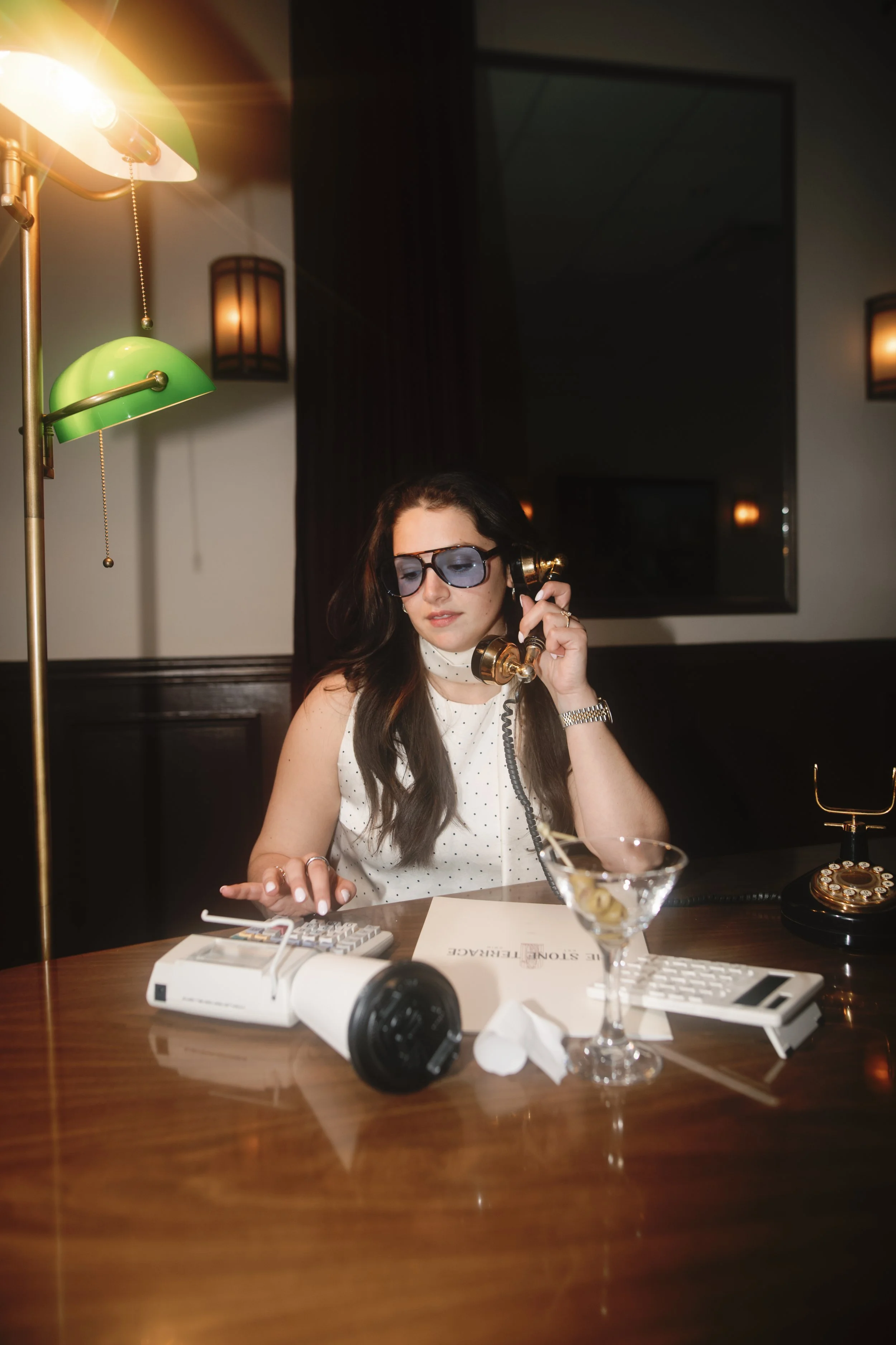 A woman wearing sunglasses, sitting at a wooden desk with a vintage telephone, calculator, and a martini glass with olives, talking on a gold-colored rotary phone in a dimly lit room.