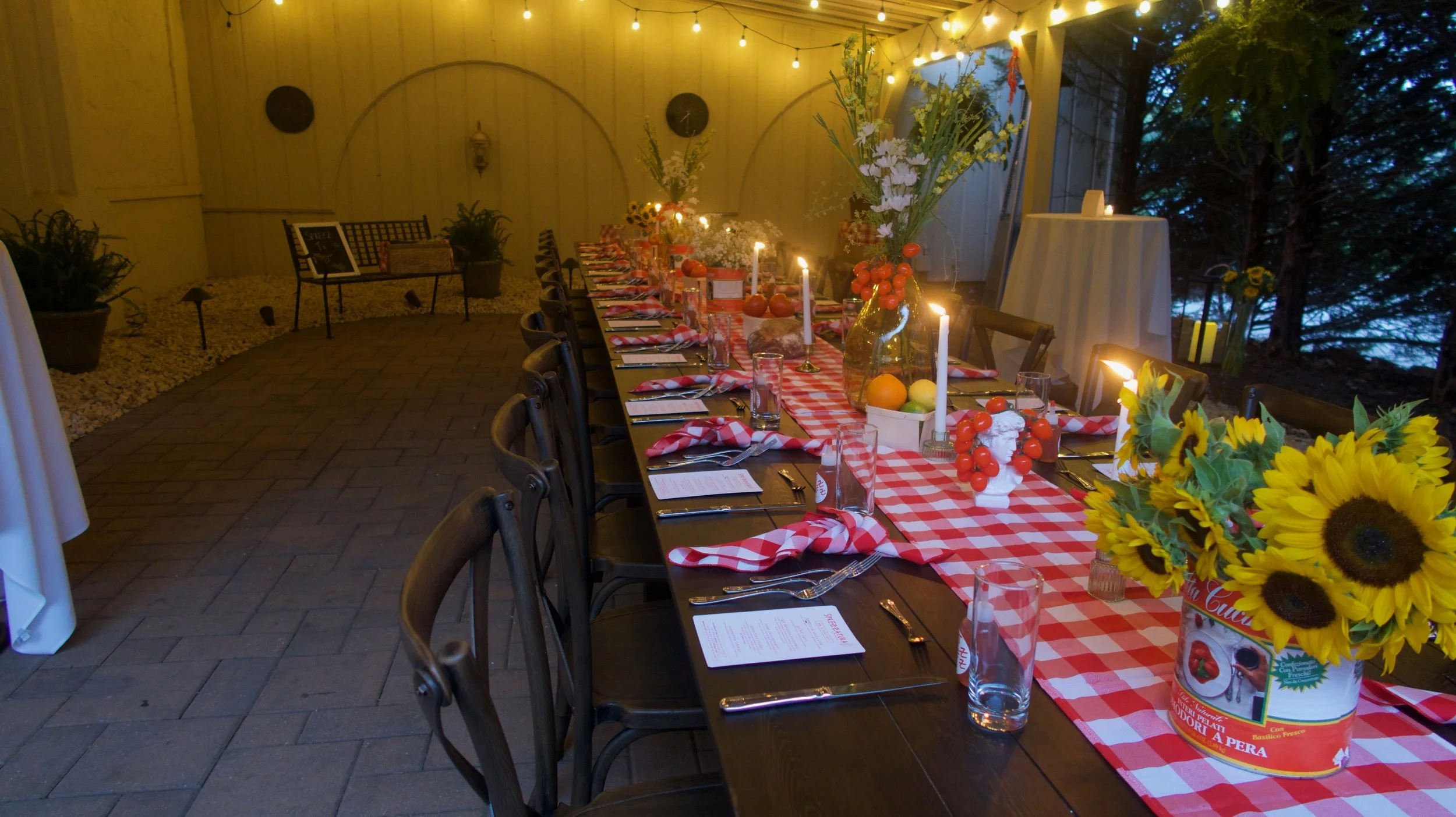Long dining table decorated with red and white checkered tablecloth, floral arrangements, candles, and eating utensils, set for a meal in a warmly lit outdoor space with string lights and greenery.