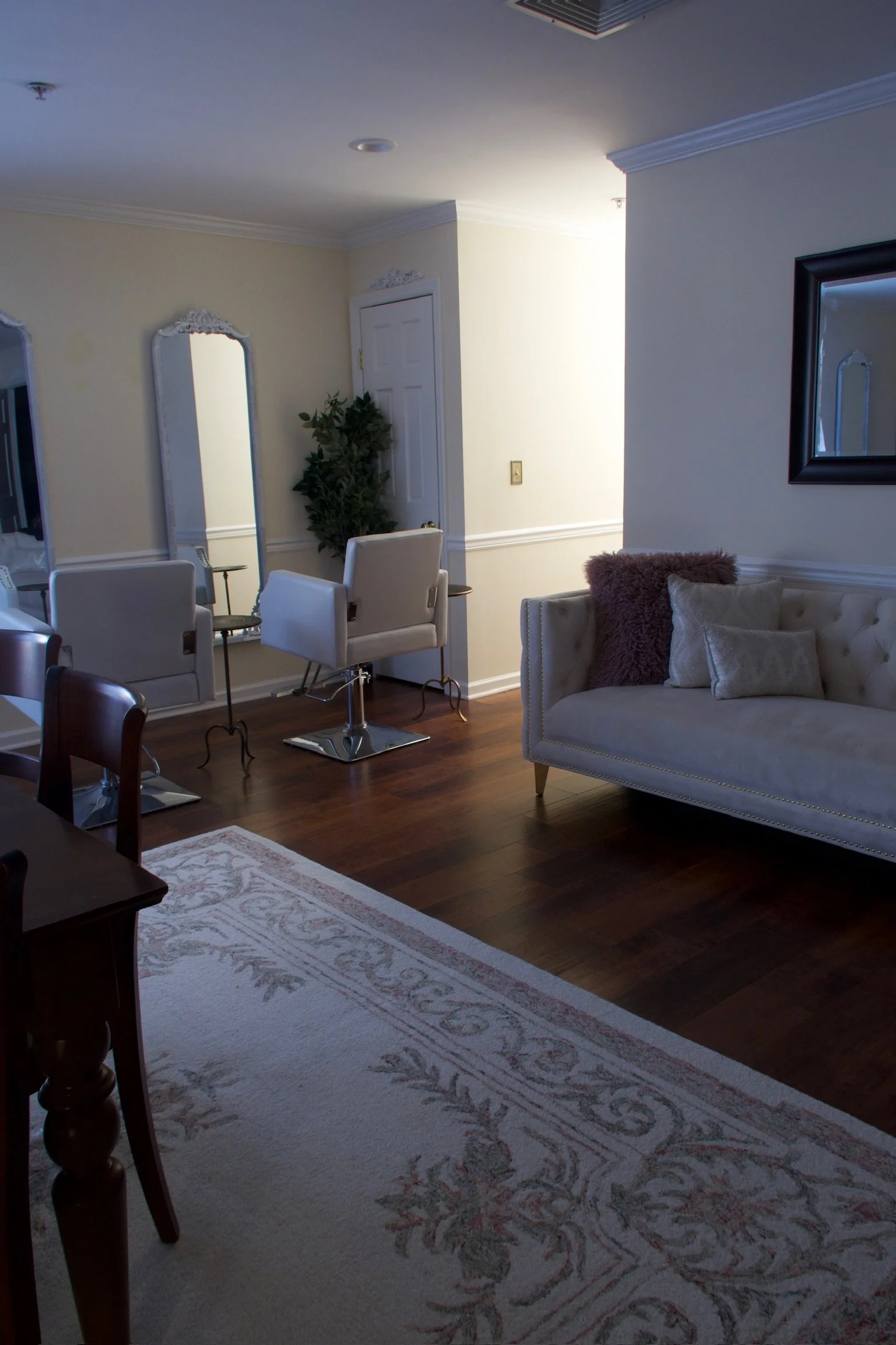 A living room with wooden flooring, a white tufted couch with pillows, a large mirror, a potted plant, two salon chairs, a side table, and a rug with floral patterns.