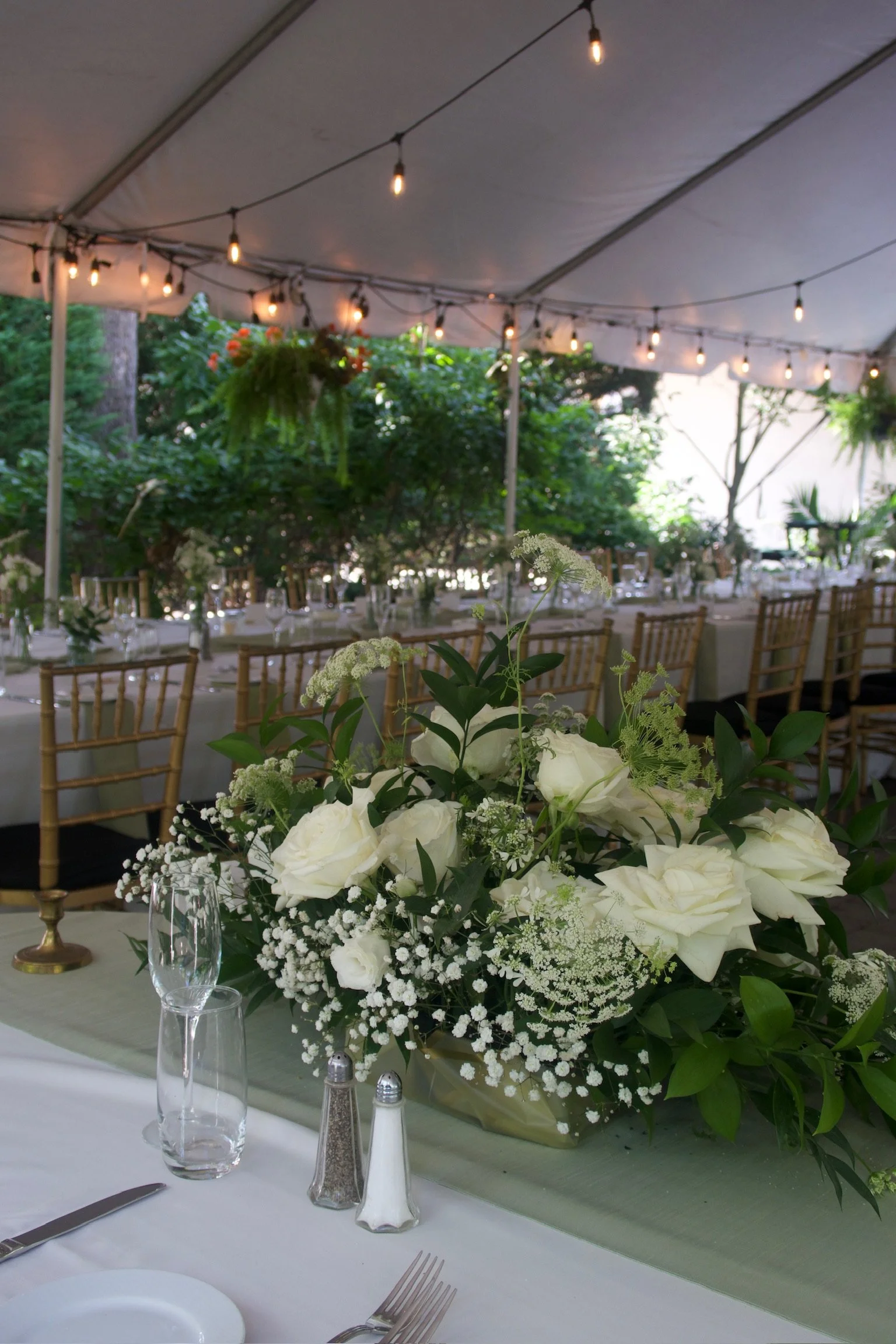 A floral centerpiece with white roses, baby's breath, and greenery on a table at an outdoor event under a tent with string lights.