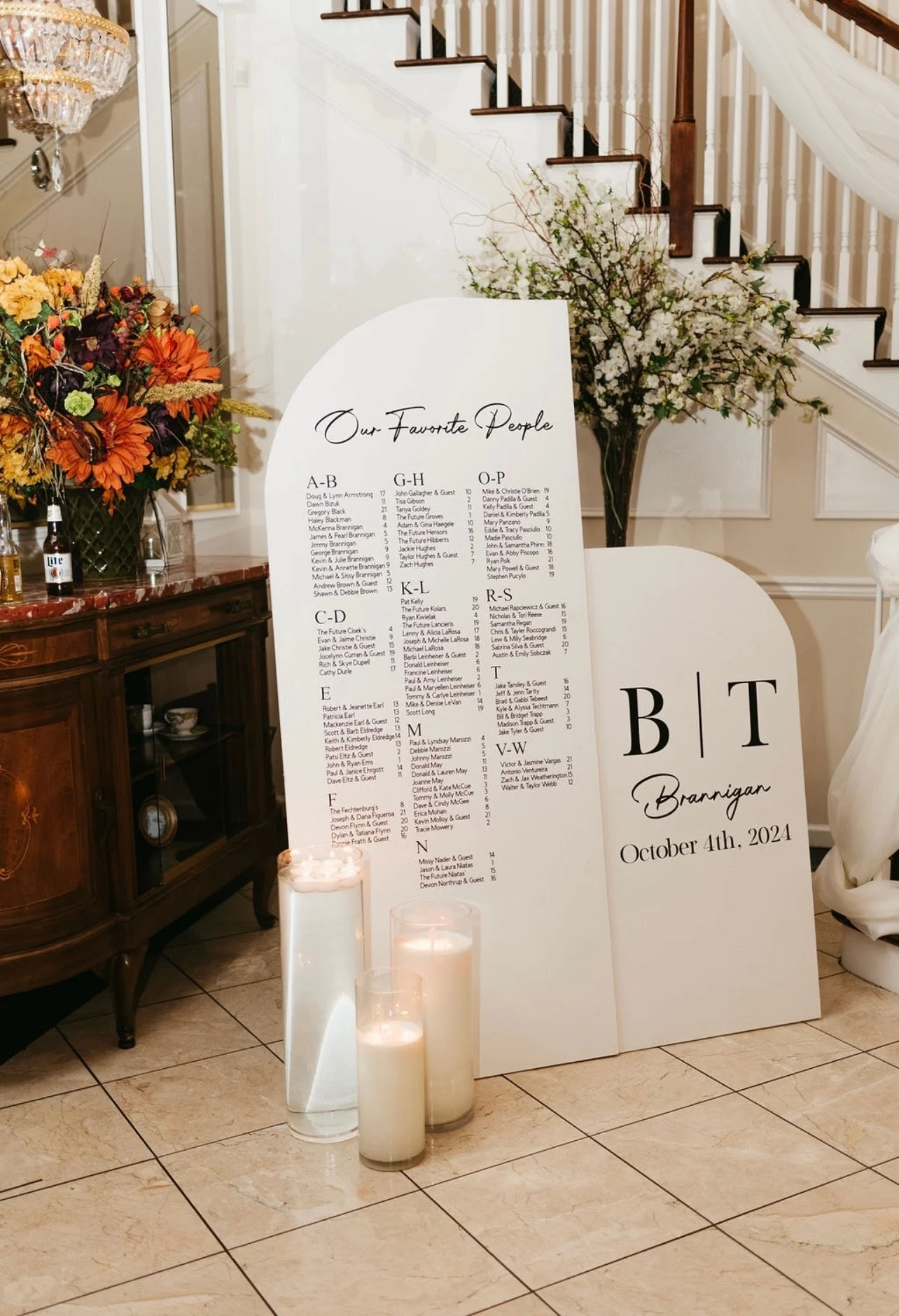 A wedding seating chart board displaying guests' names and table assignments, labeled 'Our Favorite People.' The board is decorated with floral arrangements, candles, and is situated in a room with a staircase and elegant decor.