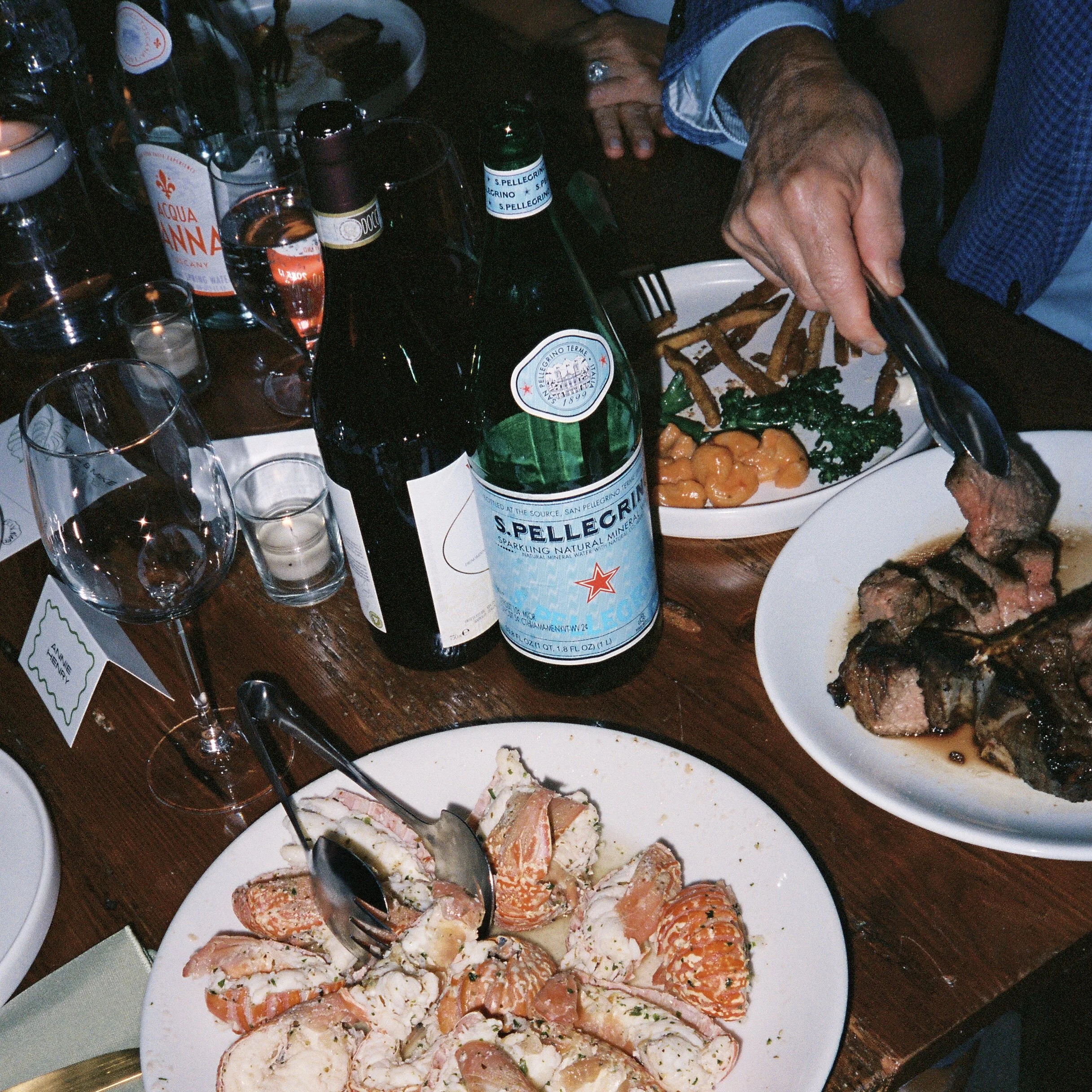 A table set with split lobster, steak, and side dishes. There are bottles of San Pellegrino sparkling water, wine glasses, and lit candles. A person is serving food with tongs.
