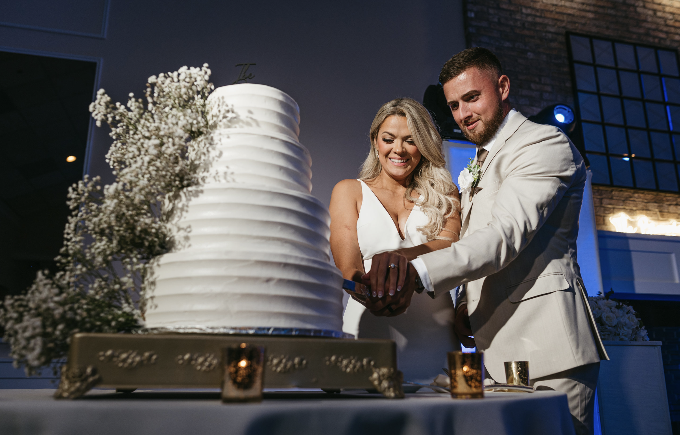 A newlywed couple in wedding attire cutting a large white wedding cake together at their reception.