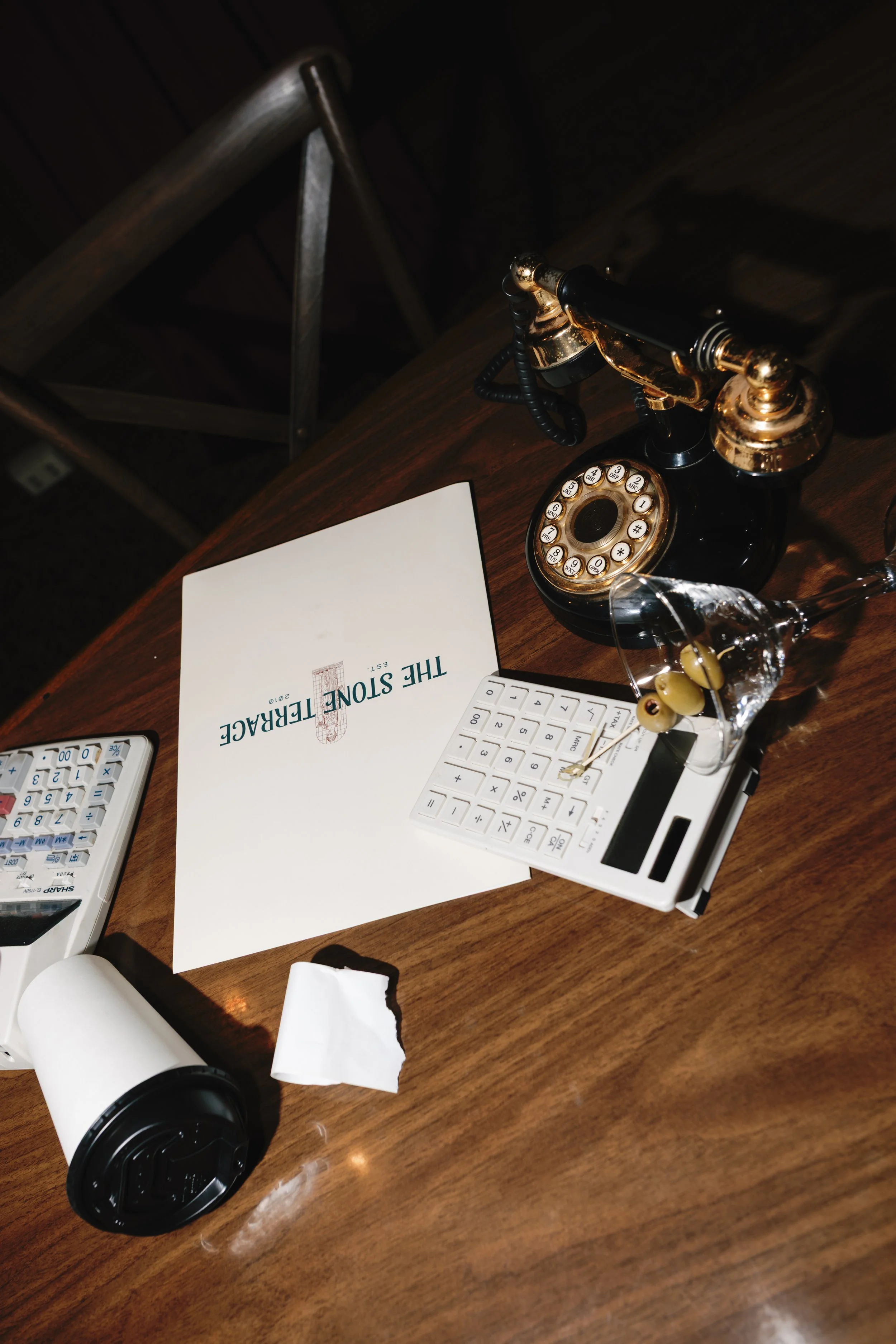 A wooden desk with vintage black and gold rotary telephone, a calculator, a glass with toothpicks and olives, a white paper cup, a receipt, and a menu titled 'The Stone Terrace'.