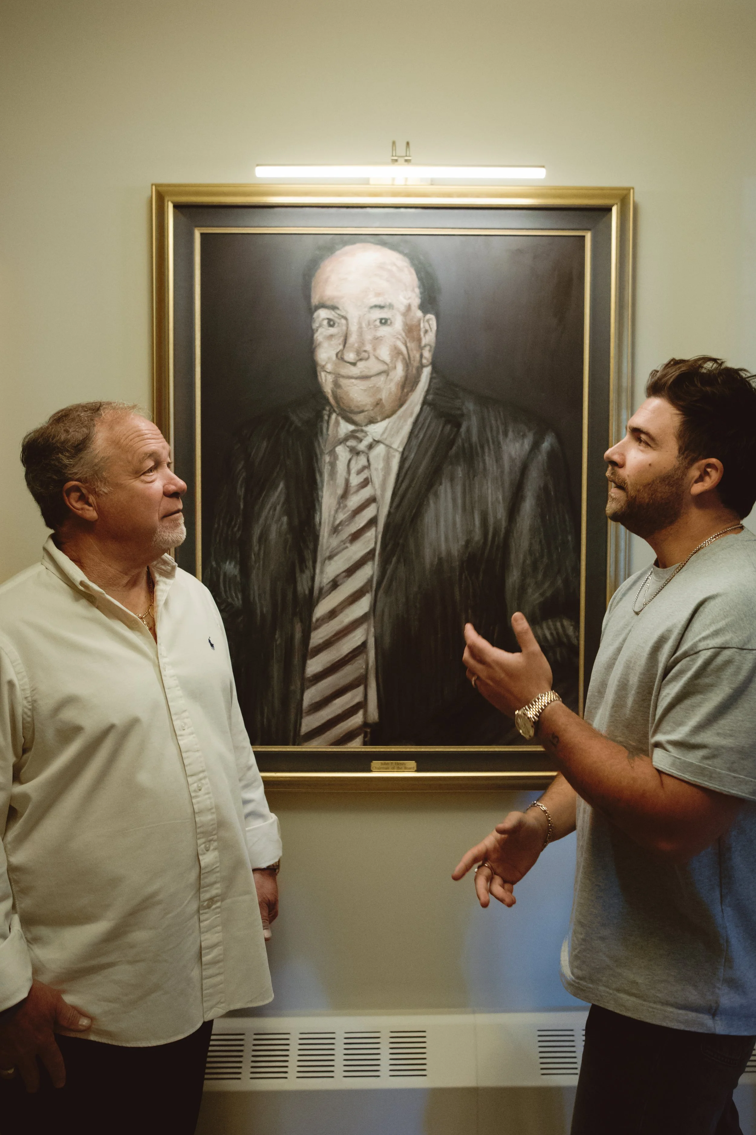 Two men are standing and talking in front of a large framed black-and-white portrait of a smiling man in a suit and tie on display in a gallery or museum setting.