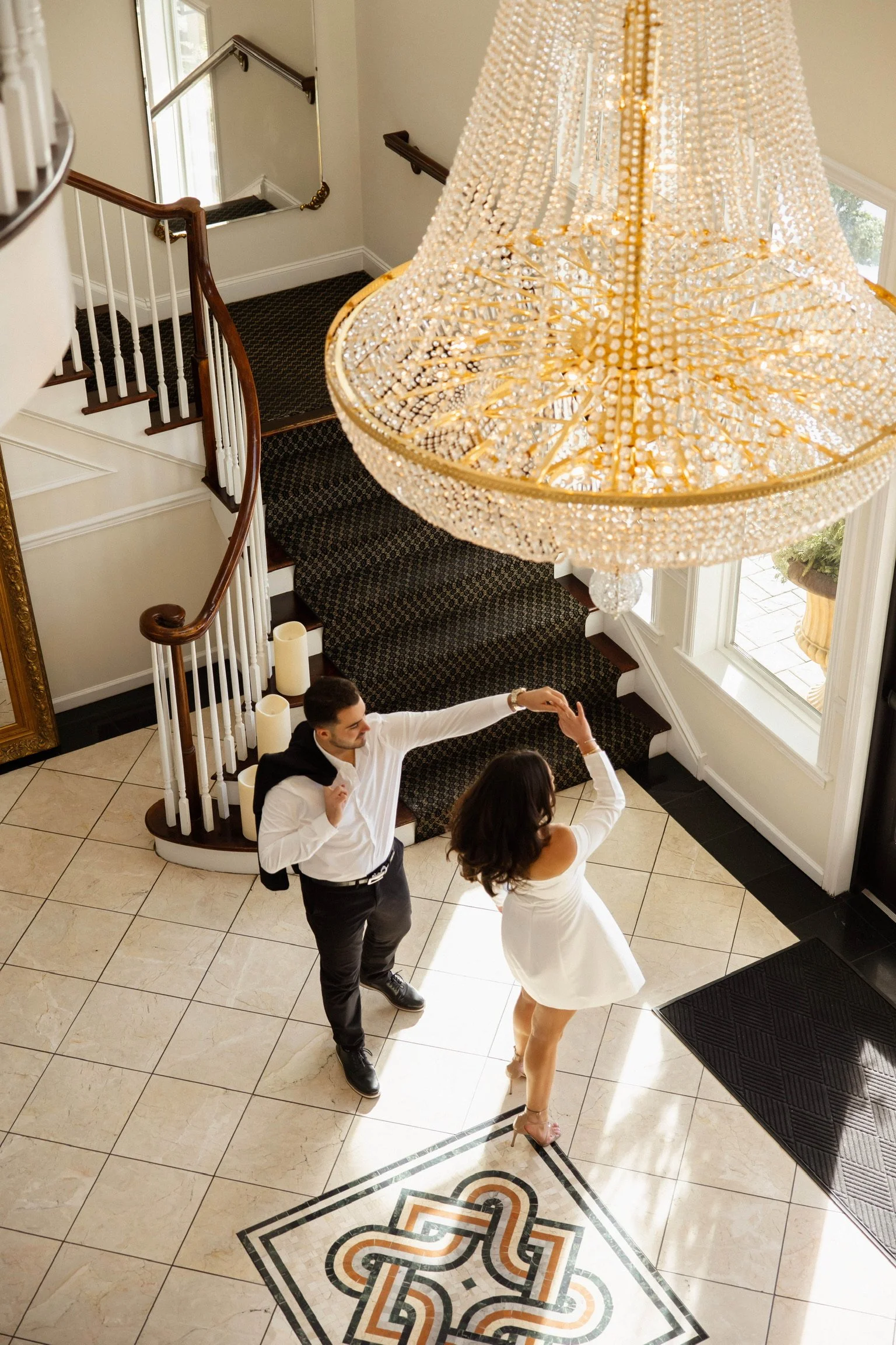 A man and woman dancing in a lobby near a staircase with a large chandelier overhead.