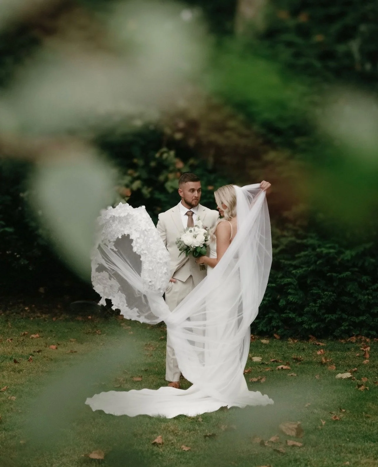 A bride and groom standing outdoors in a garden, with the bride holding a bouquet of white flowers and the groom dressed in a light-colored suit. Green foliage in the background and leaves on the grass.