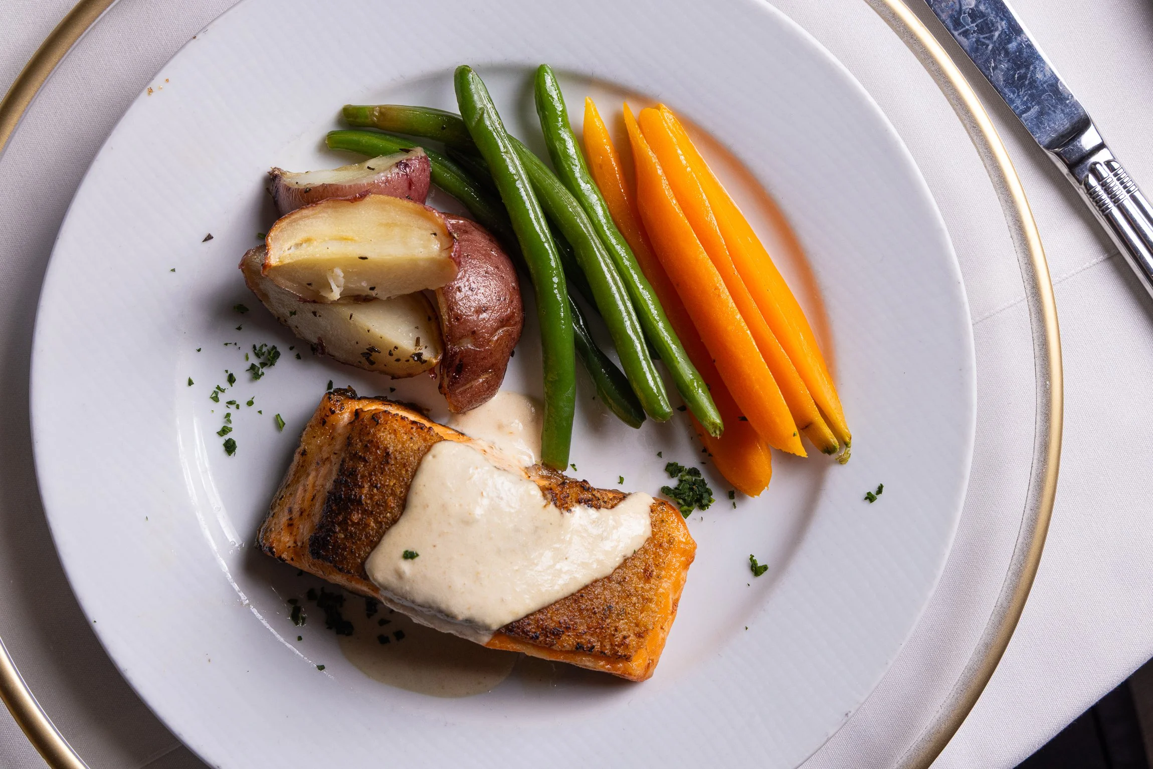 A plate with grilled salmon topped with white sauce, roasted potatoes, green beans, and sliced carrots, garnished with chopped herbs.