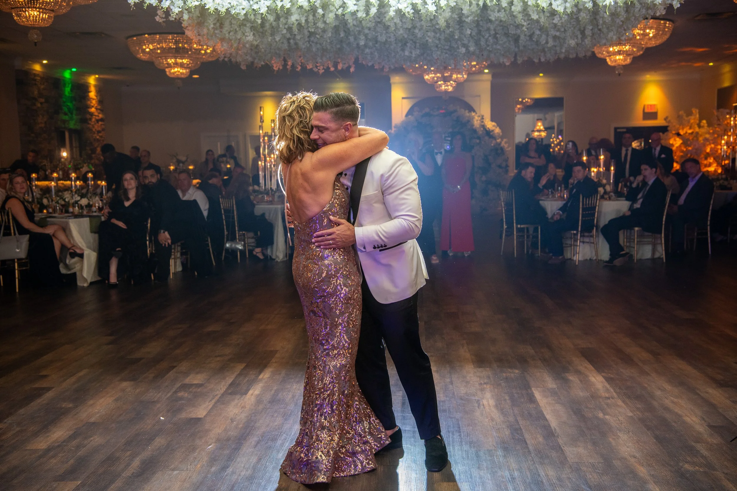 A man and woman dancing closely on a dance floor at a wedding reception, surrounded by seated guests in an elegant hall decorated with flowers and chandeliers.