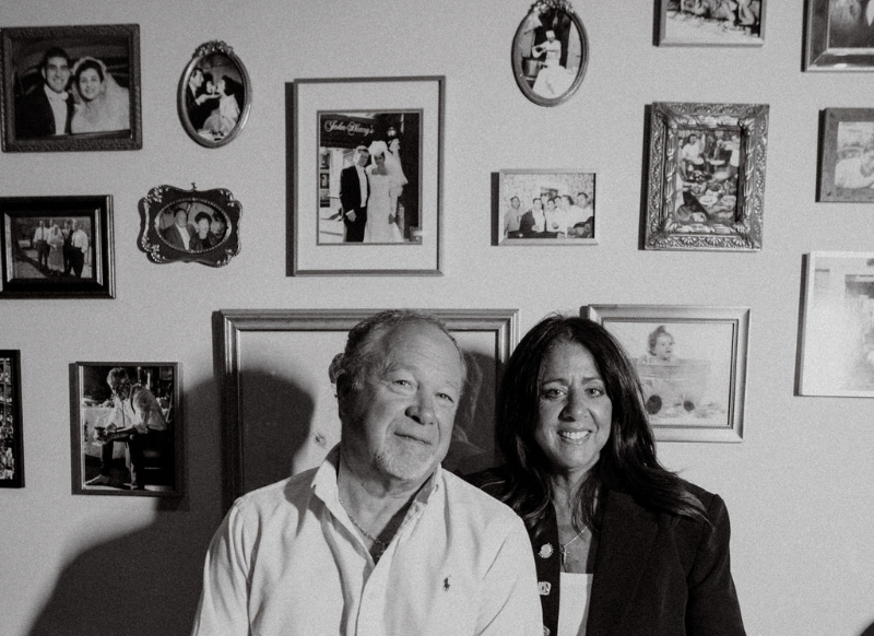 A black-and-white photo of an older man and a woman smiling and sitting close together in front of a wall decorated with various framed photographs.