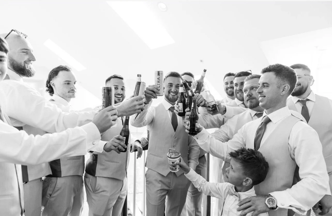 Group of men and a boy in formal attire raising drinks in celebration, smiling, indoors with white ceiling and skylights.