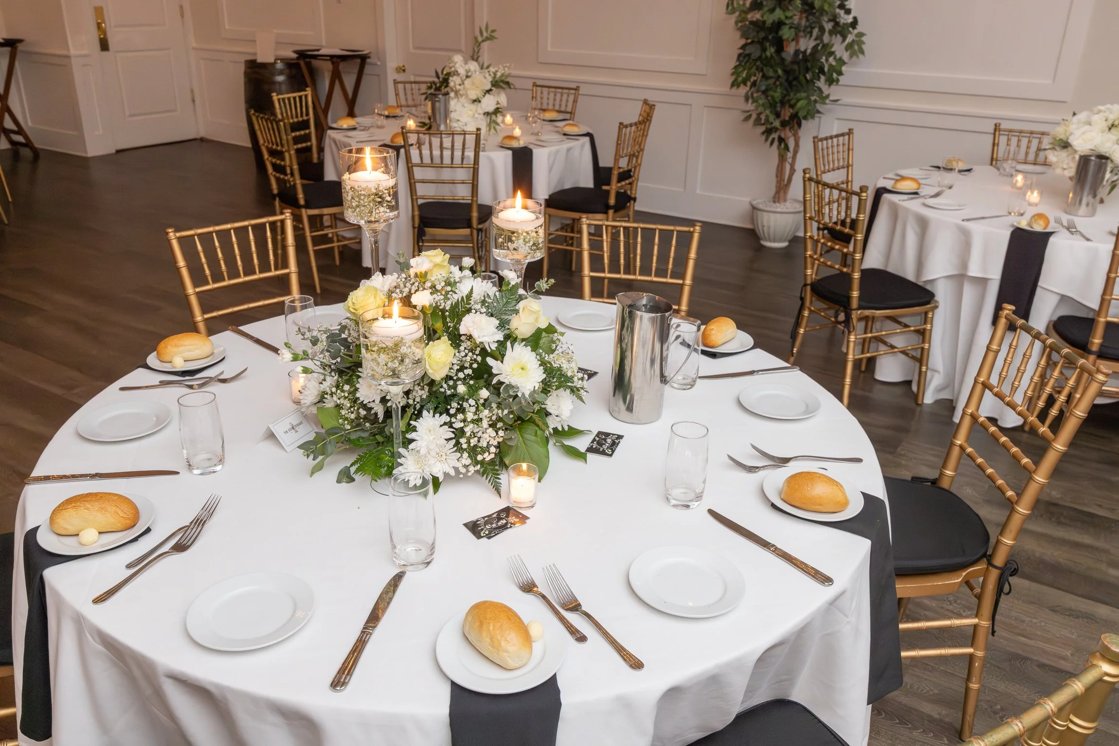 Round banquet table set for a formal event with white tablecloths, floral centerpiece, candles, bread rolls, and place settings including plates, silverware, and glasses, surrounded by gold chairs with black cushions in a decorated room.