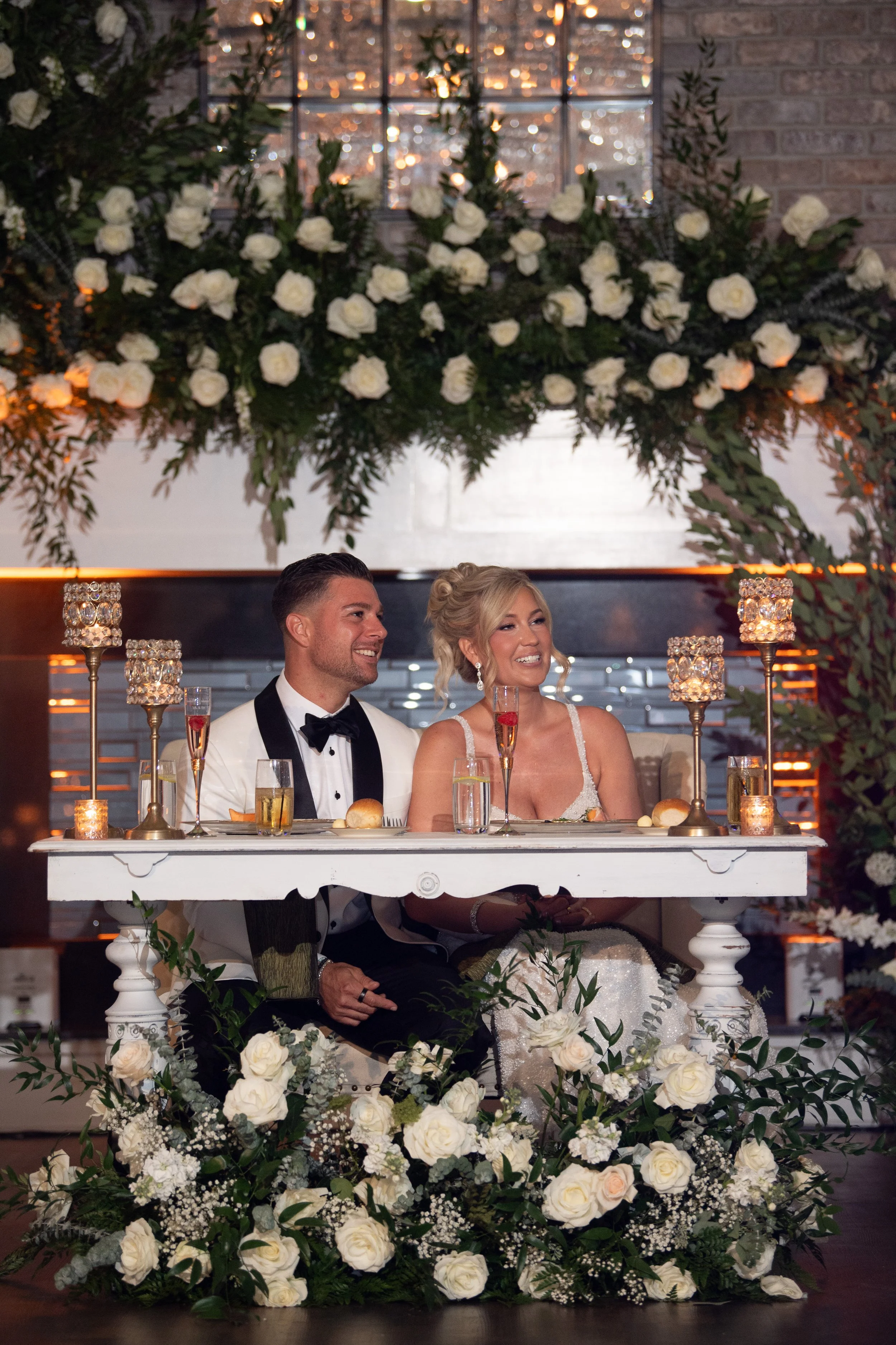 A bride and groom sitting at a decorated head table during their wedding reception, with floral arrangements and candleholders, smiling and enjoying their moment.