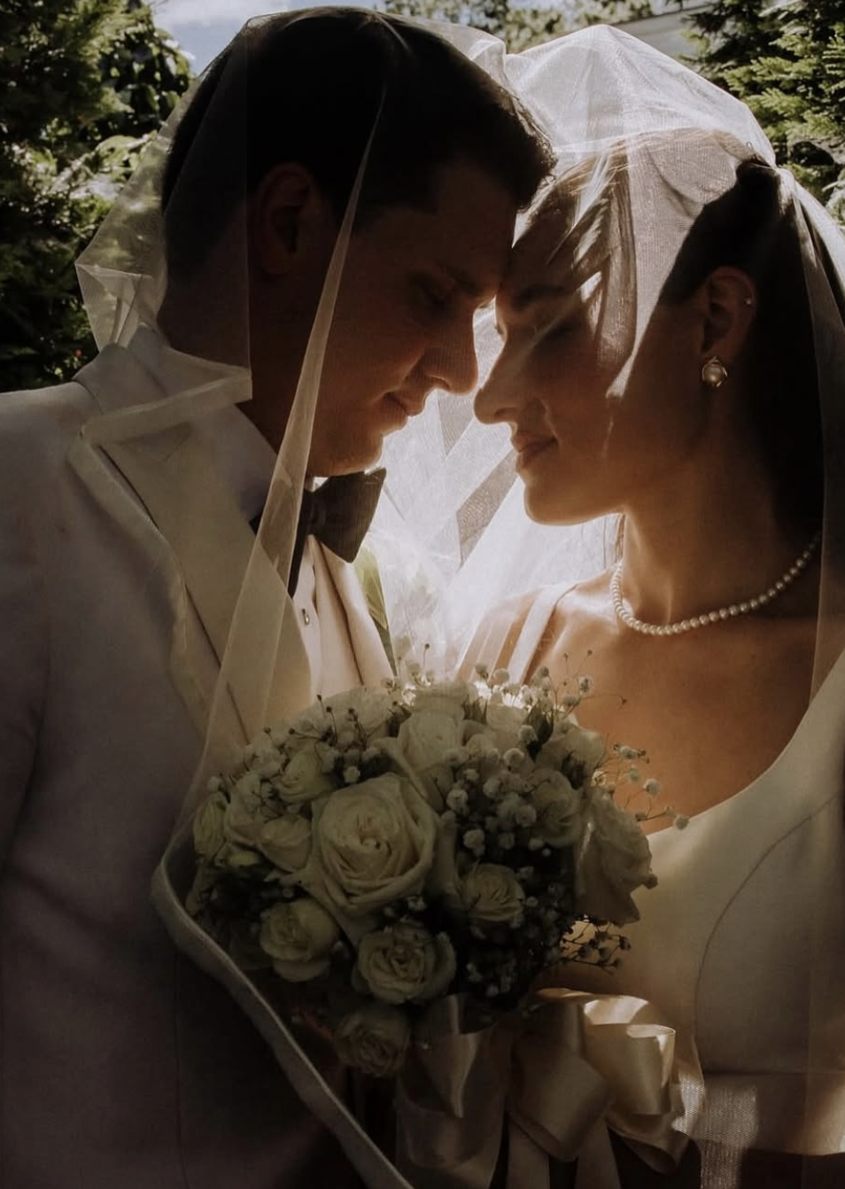 A bride and groom on their wedding day, close-up, with their foreheads touching, holding a bouquet of white roses and baby's breath, wearing wedding attire and veils.
