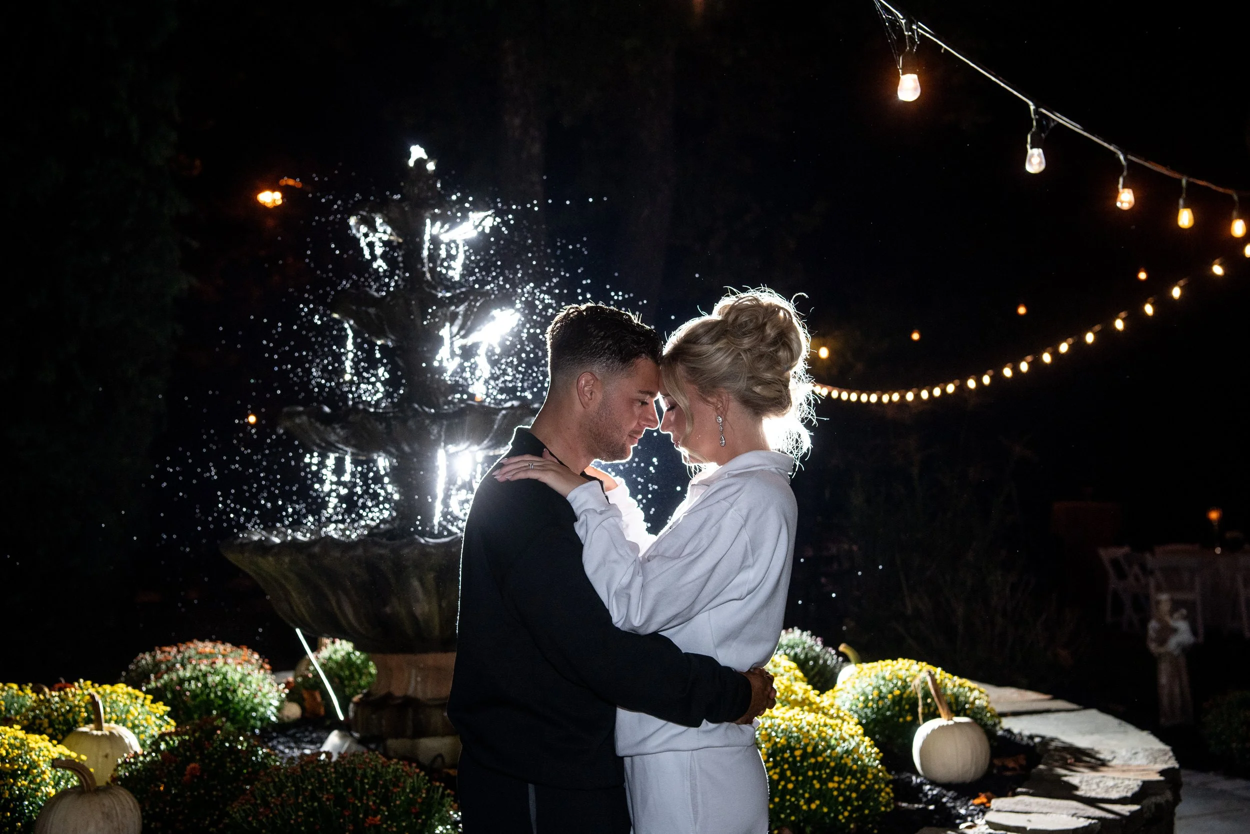 A romantic couple stands closely together during nighttime, with their foreheads touching and eyes closed. They are illuminated from behind, in front of a large fountain with water splashing, surrounded by pumpkins and flowers, under string lights.