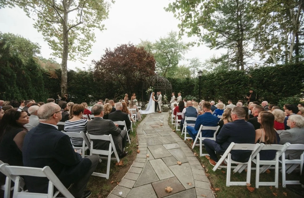Outdoor wedding ceremony with bride and groom standing in front of officiant, seated guests on both sides, garden setting, trees and bushes surrounding, overcast sky.