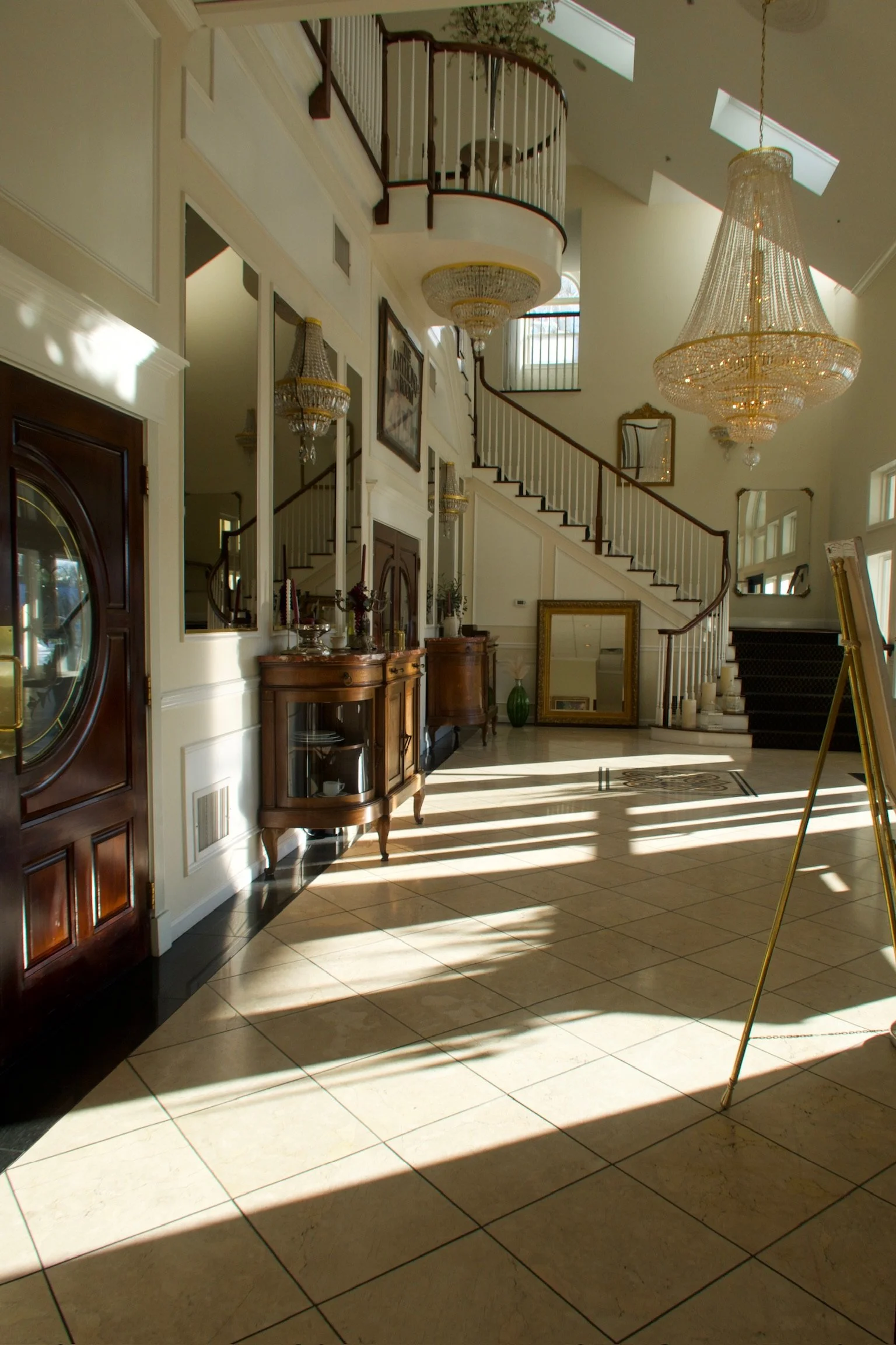 Elegant hotel lobby with grand staircase, large chandeliers, and sunlight streaming through windows, creating striped shadows on the tiled floor.