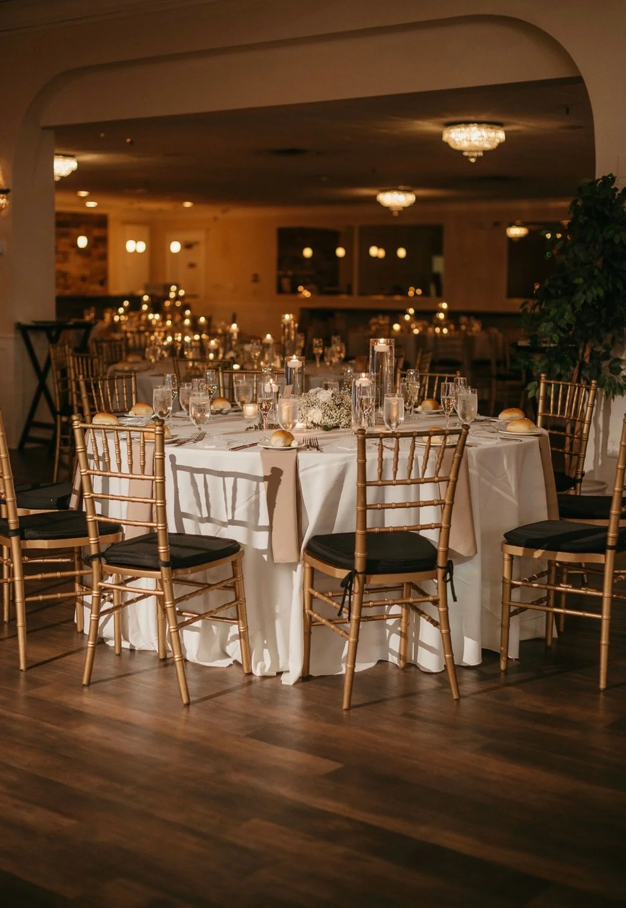 Elegant banquet table set for a formal event, with gold chairs, white tablecloth, glassware, candles, and a flower centerpiece in a dimly lit room.