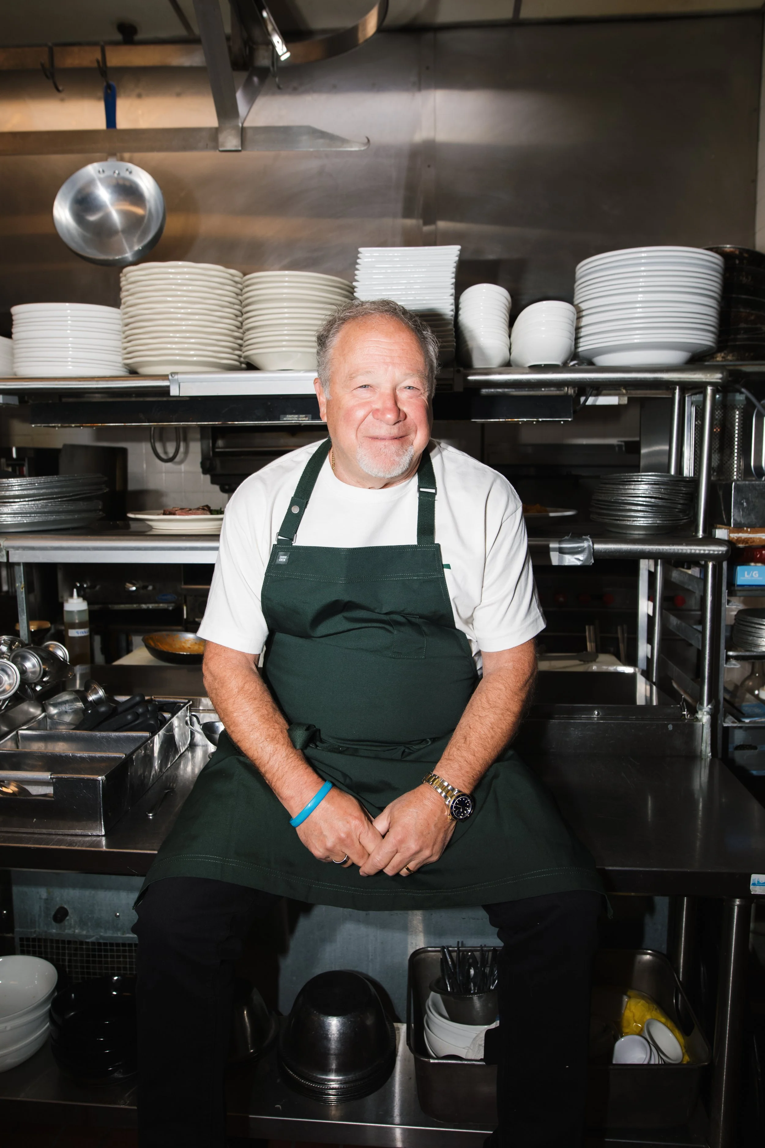 A man sitting in a professional kitchen, wearing a dark green apron and a white shirt, surrounded by stacked white and black plates, with kitchen utensils and equipment nearby.