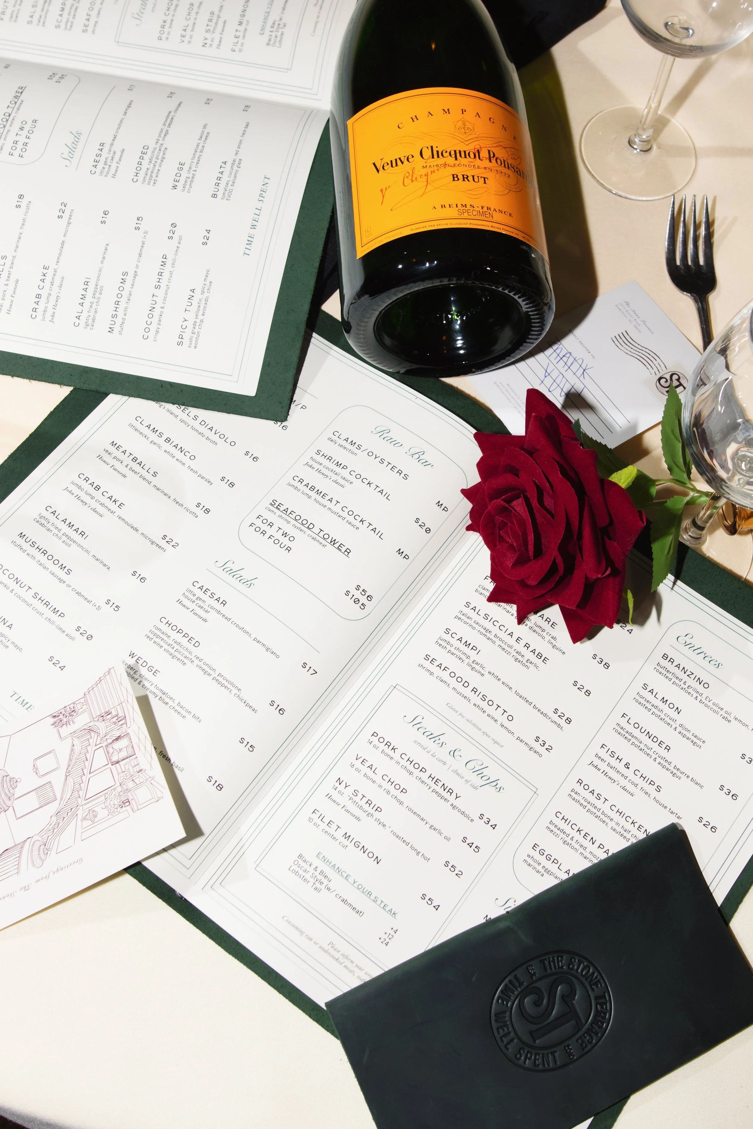 A table setup with a menu, a bottle of Veuve Clicquot Champagne, a glass of water, a fork, and a red rose.
