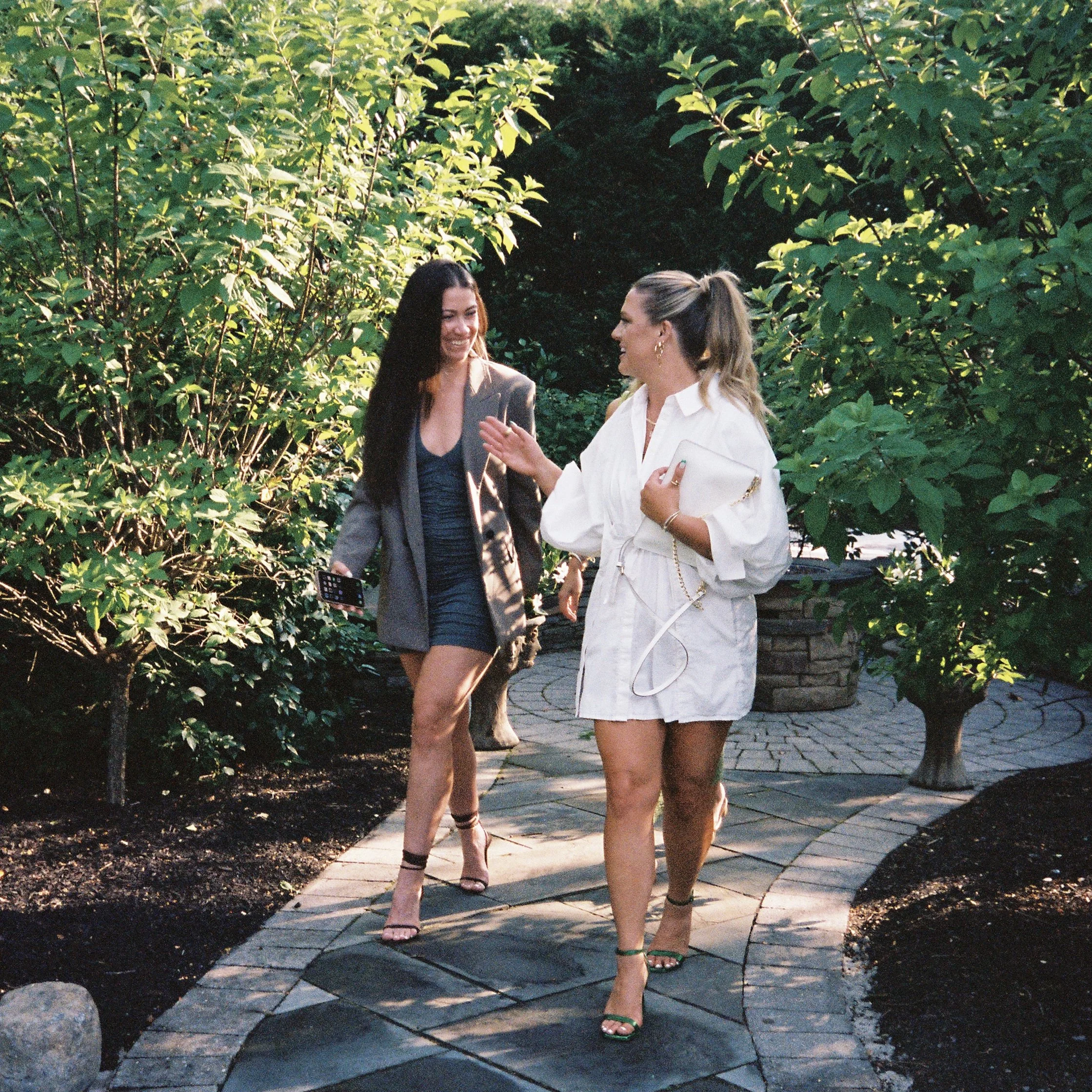 Two women walking and talking on a garden path surrounded by lush green bushes.