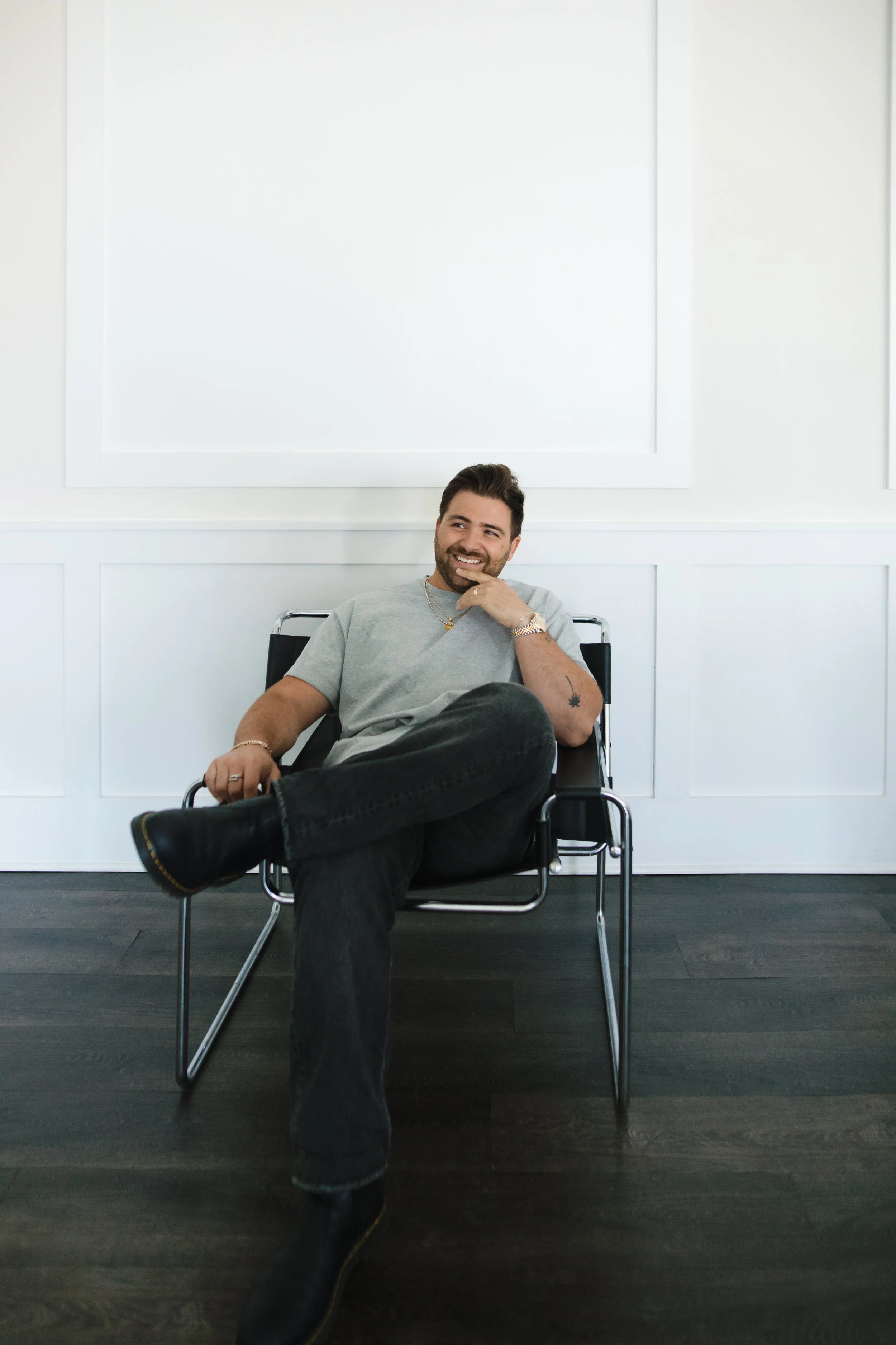 A man with a beard and tattoos, wearing a light gray t-shirt, black jeans, and black boots, sitting in a modern chair with one leg crossed over the other, smiling and looking to his right in a minimalistic room with white walls and dark wooden flooring.