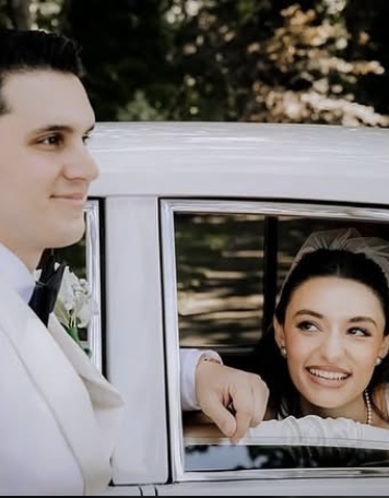 A bride and groom, dressed in wedding attire, on a wedding day, with the bride smiling and looking at the groom, who is standing next to a vintage car outdoors.