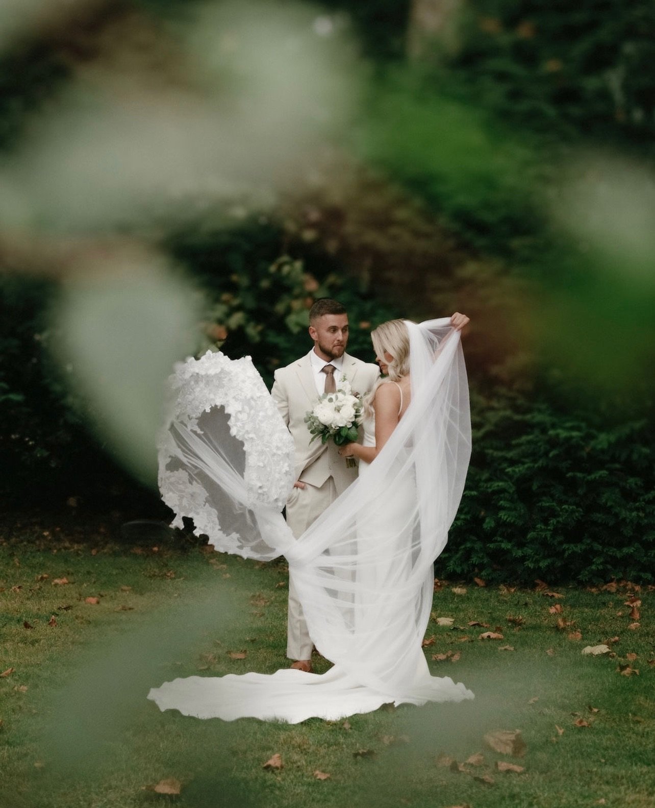 A bride and groom in wedding attire standing outdoors amidst greenery, with the bride holding a bouquet and her veil flowing around her.