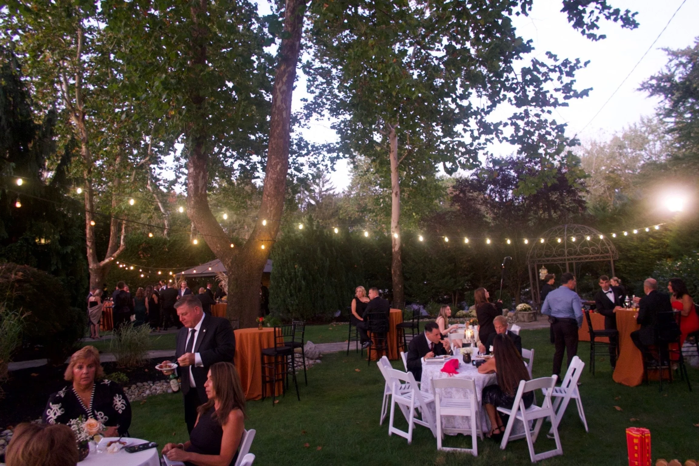 Outdoor evening gathering with guests at a garden party or wedding reception, decorated with string lights and tables with white and orange tablecloths, guests mingling and eating.