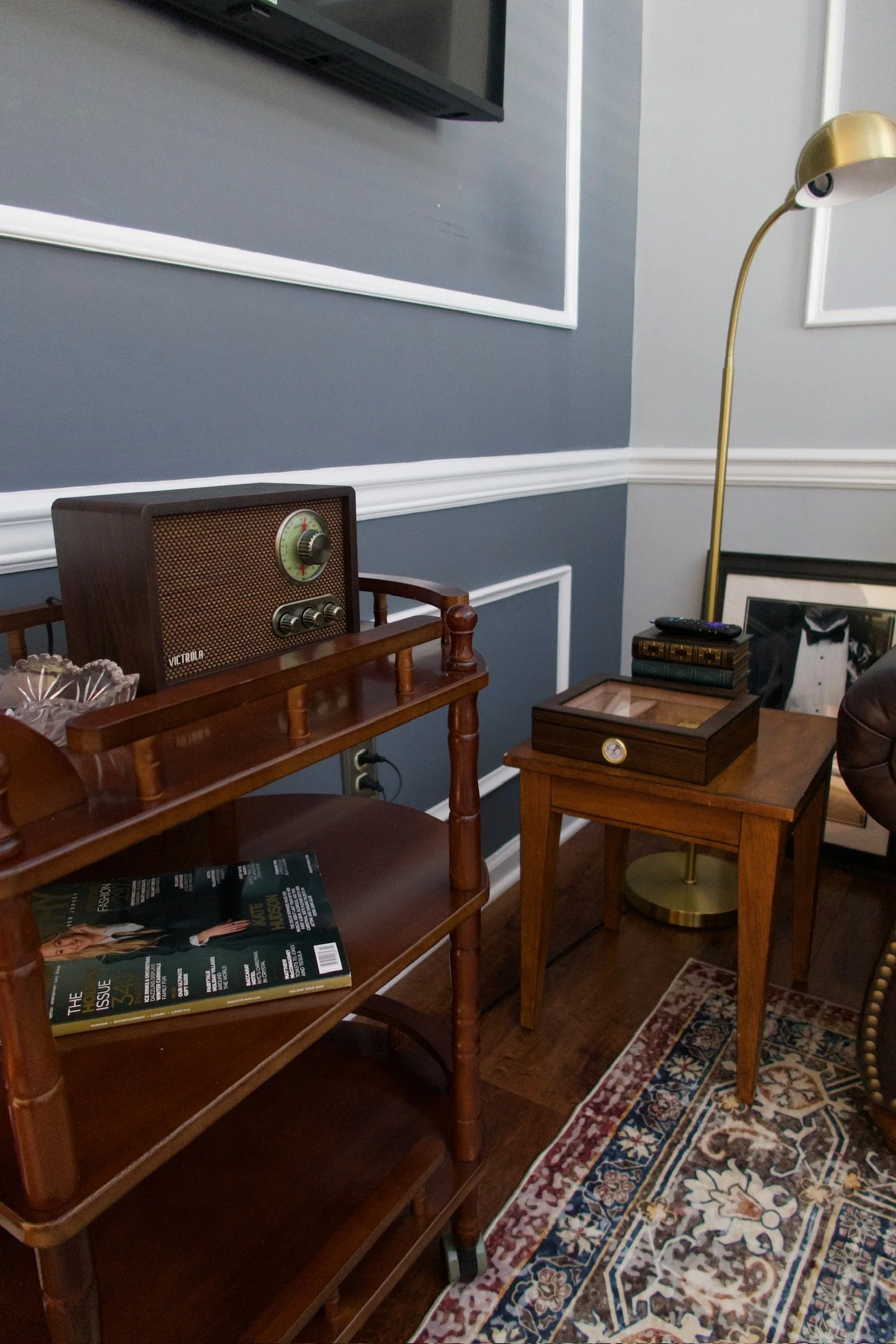 Living room corner with vintage wooden radio on a wooden cart, small wooden side table with a glass display case, a lamp, and framed artwork on the wall.