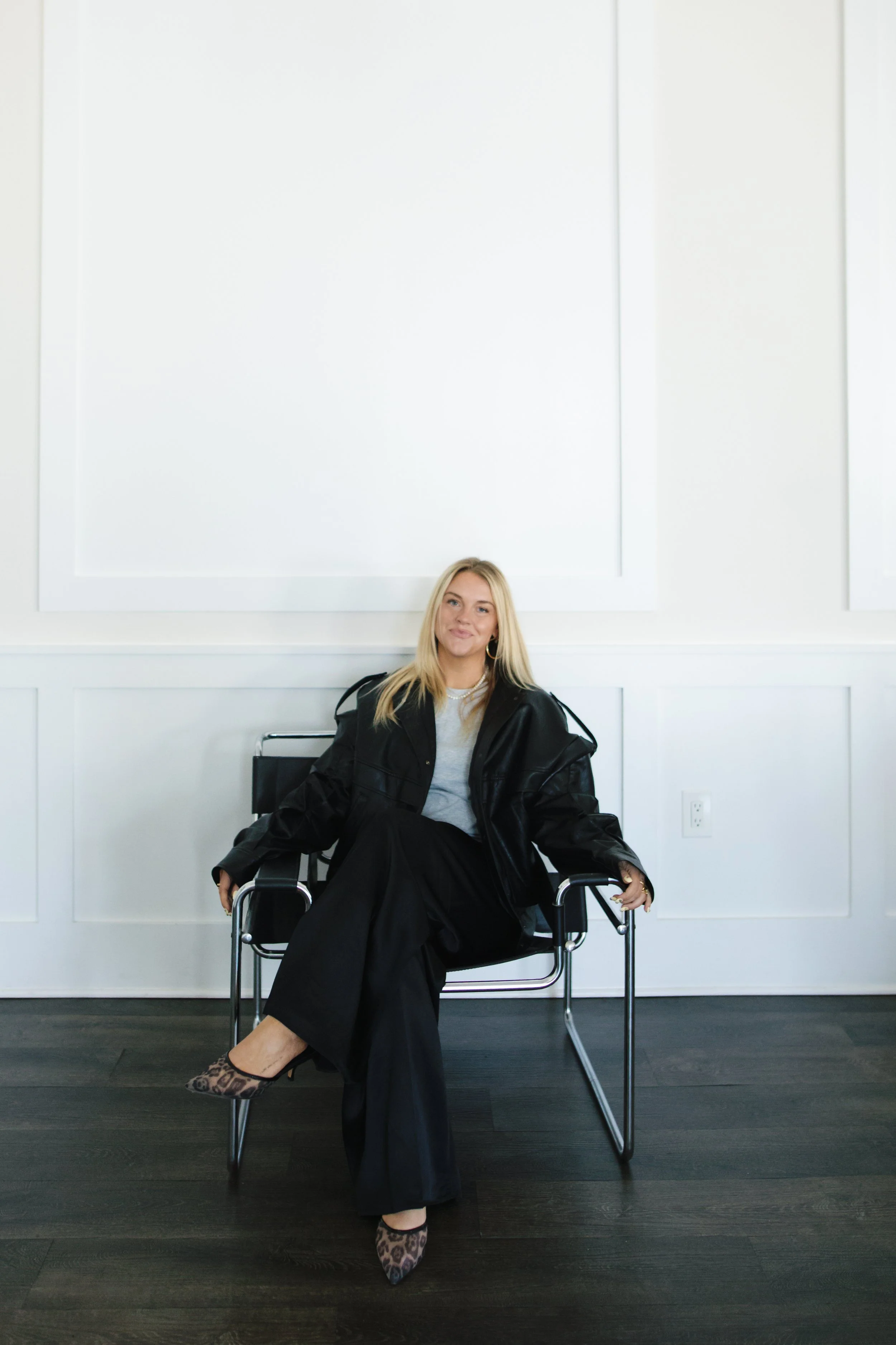 A woman with blonde hair sitting on a black chair in a minimalist room with white walls and dark wooden flooring, wearing leopard print shoes, a gray top, black pants, and a black jacket.