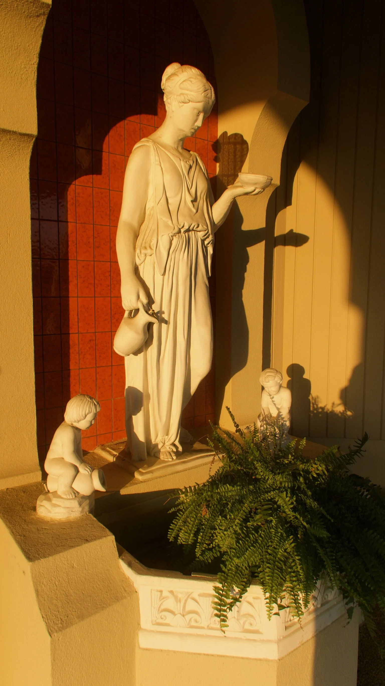 A religious statue of a woman holding a bowl in one hand and a jug in the other, with two smaller children statues at her feet, surrounded by greenery, set against a tiled wall in warm sunlight.
