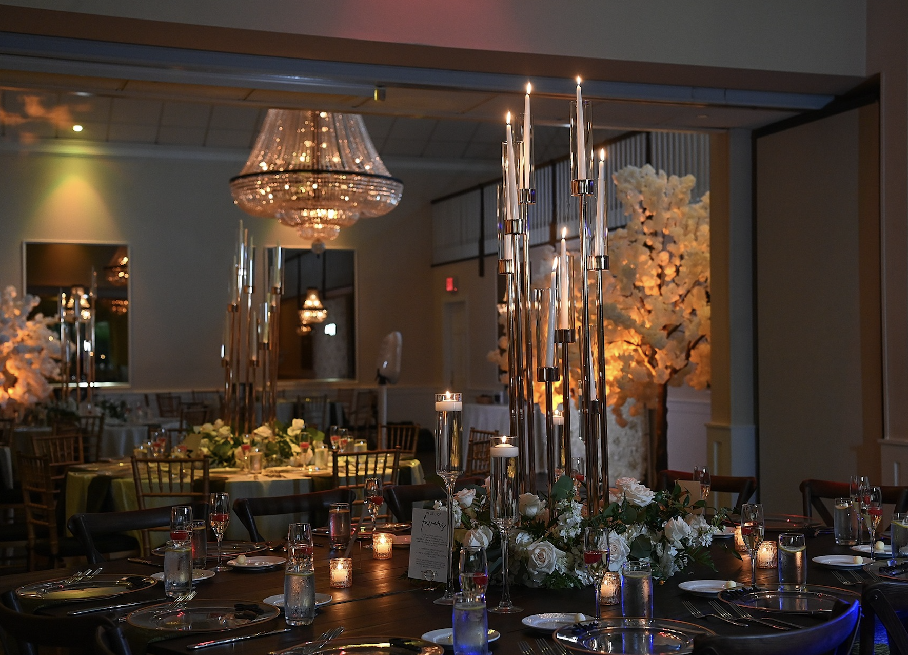 Elegant banquet table decorated with tall glass candle holders, white roses, and smaller candles, with a large chandelier hanging from the ceiling and white floral trees in the background.