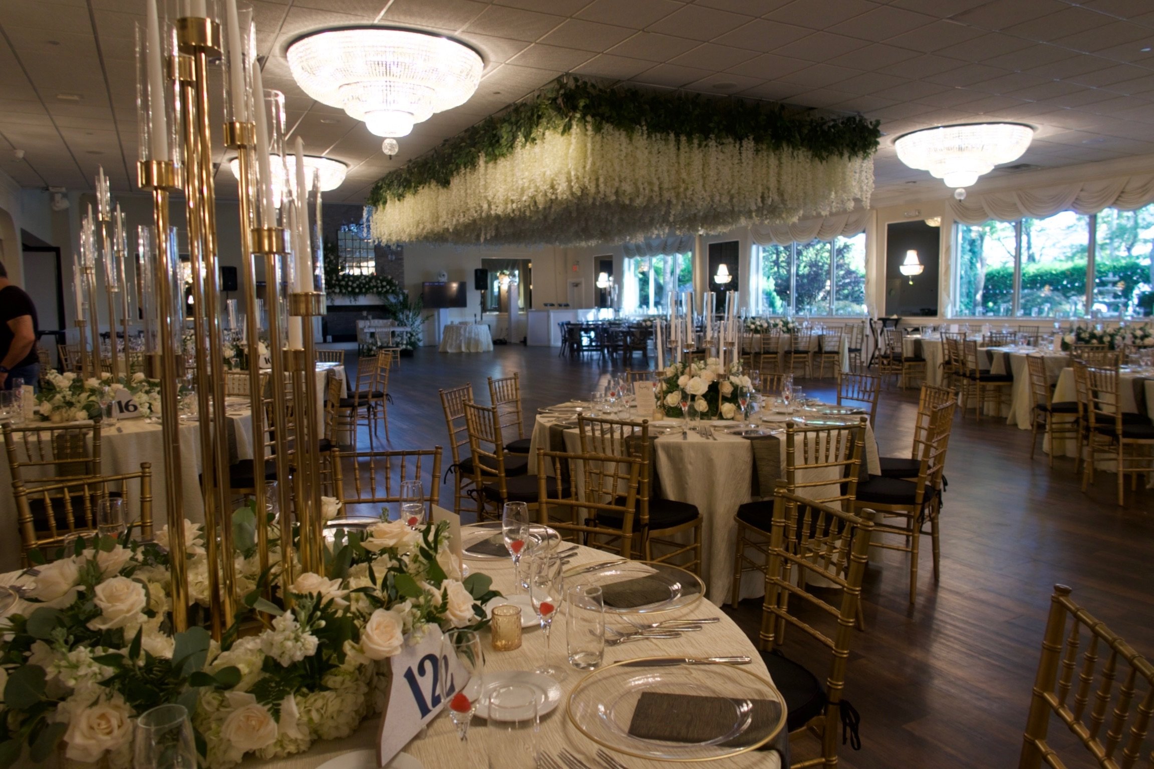 Elegantly decorated wedding reception hall with round tables covered in white tablecloths, gold chairs, floral centerpieces, and candle holders. Large floral arrangements and chandeliers decorate the ceiling, with large windows allowing natural light