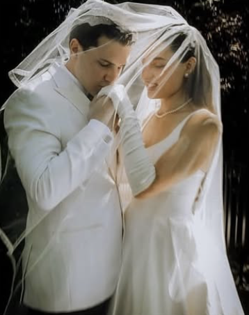 A bride and groom, dressed in white, under a wedding veil, sharing an intimate moment outdoors.
