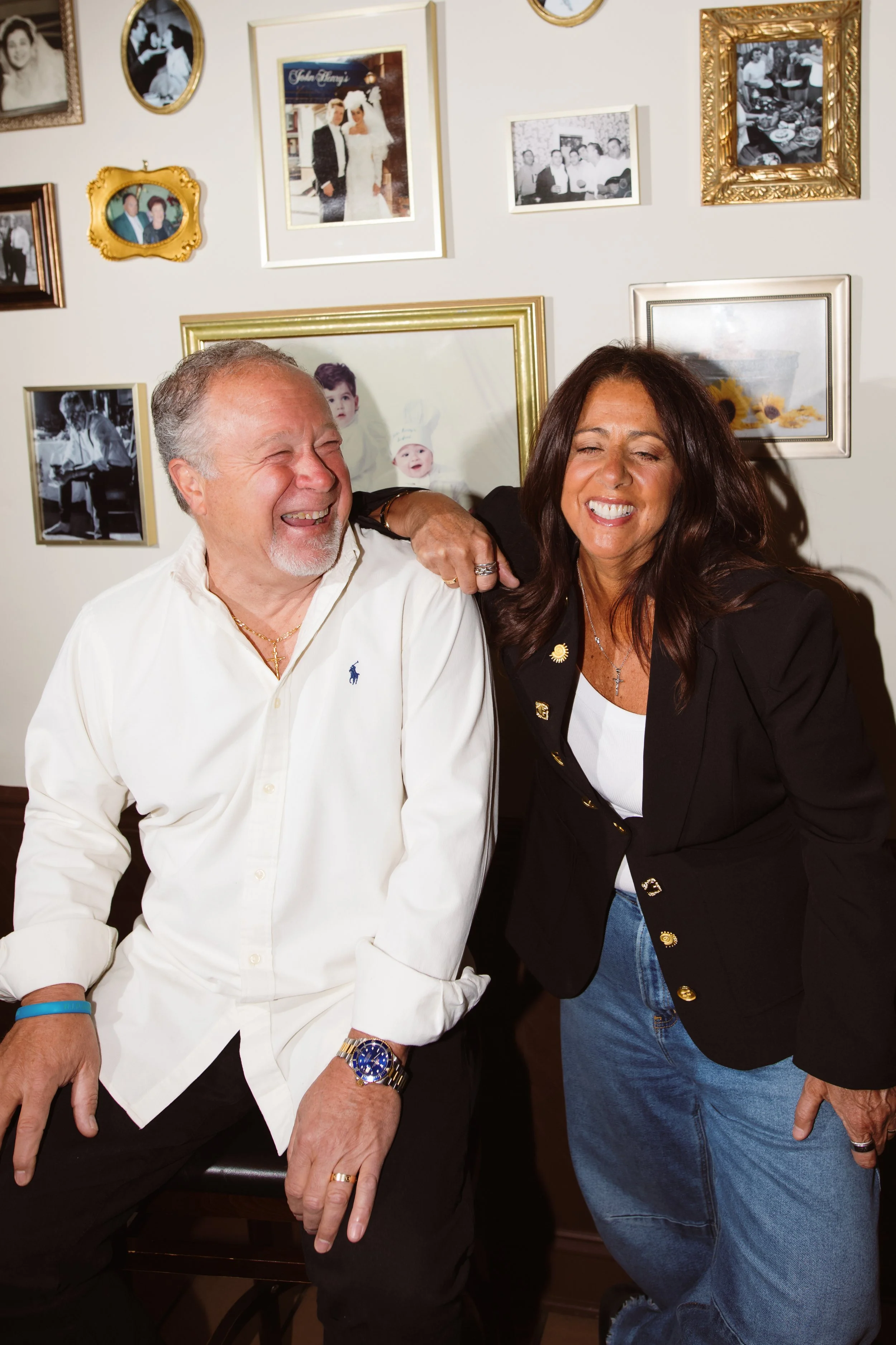 Two adults, a man and a woman, laughing and smiling in a room with a wall decorated with various framed photographs.