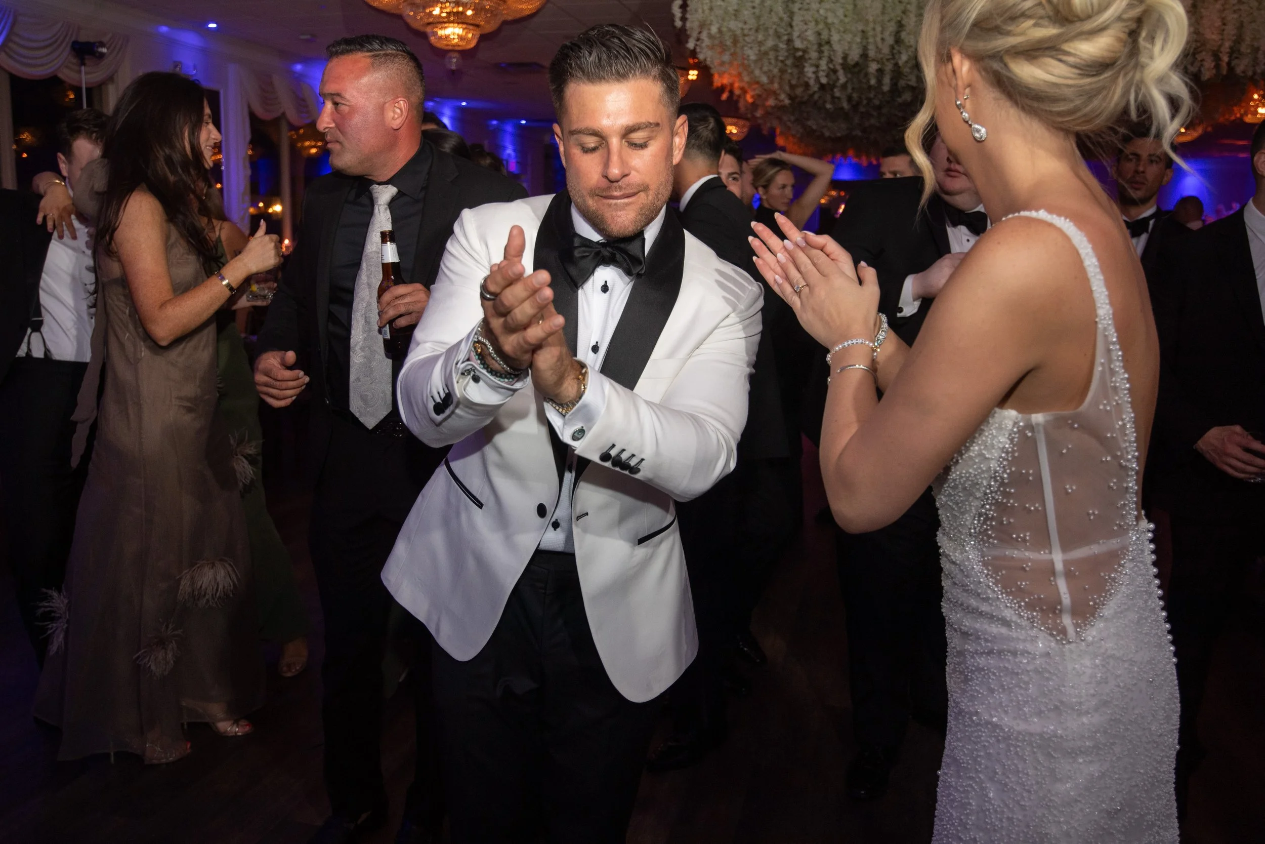 A man in a white tuxedo with black lapels and a woman in a white wedding dress with pearl embellishments dancing at a wedding reception, surrounded by other guests in formal attire in a decorated reception hall.