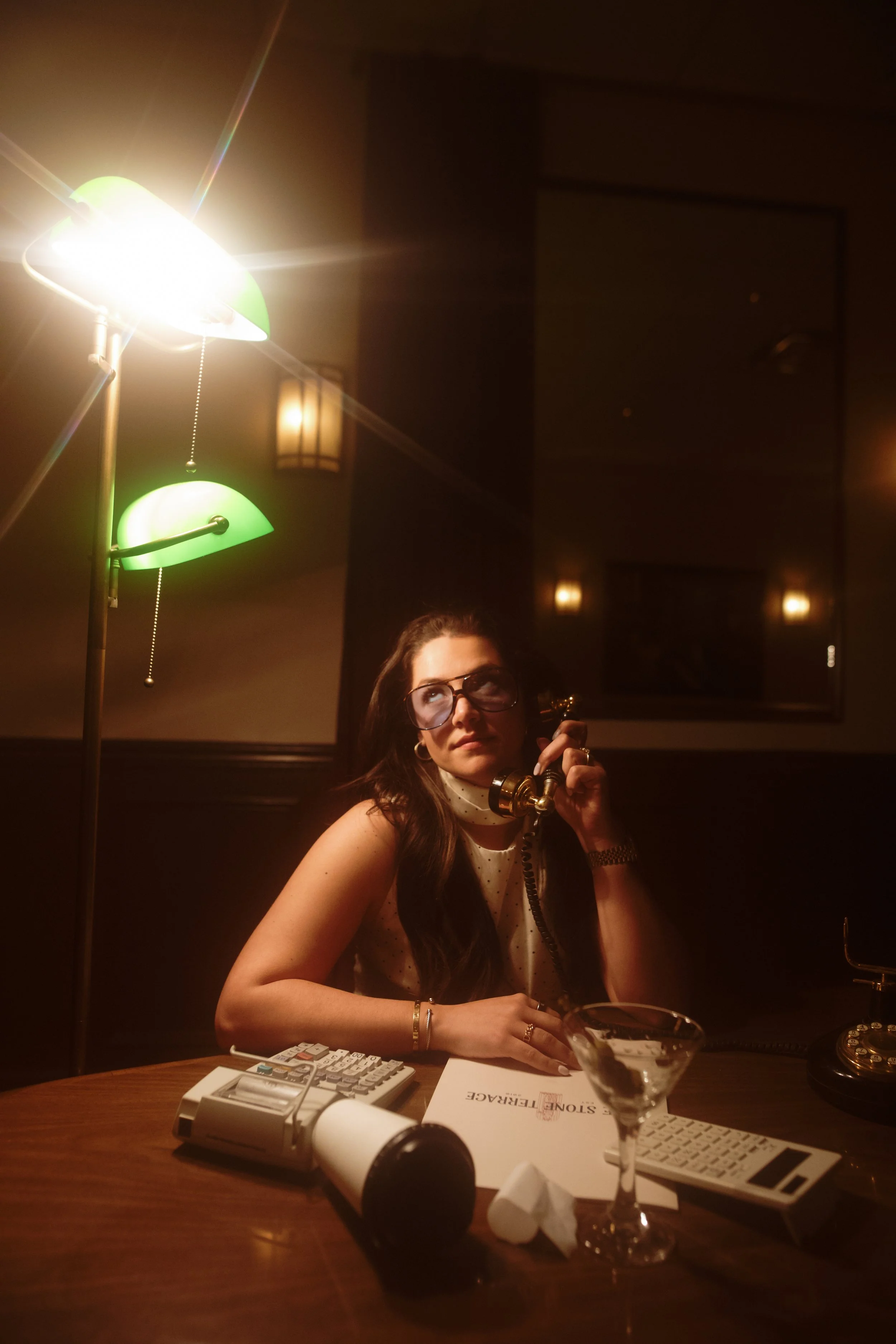 A woman with dark hair, glasses, and jewelry sits at a cluttered wooden desk with vintage music-themed decorations in a dimly lit room, talking on a rotary phone, with a cocktail glass in front of her and a calculator, remote control, and paper on th