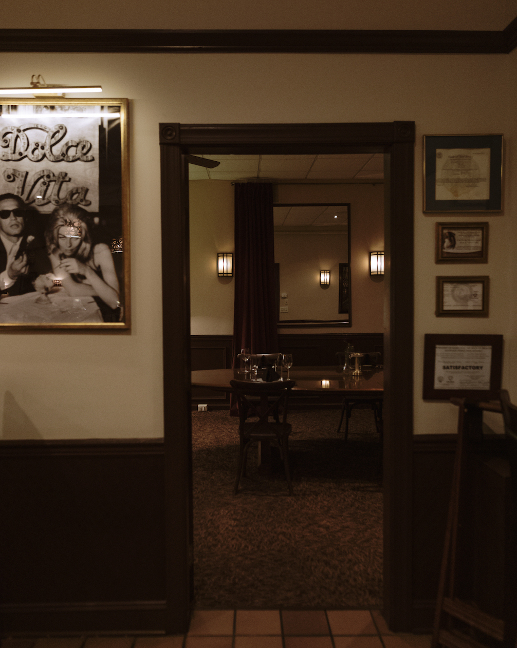A view through a doorway into a dimly lit restaurant or dining area with a wooden table set with glassware and chairs. The walls are decorated with framed artwork and certificates, and there are wall-mounted lights creating a cozy ambiance.