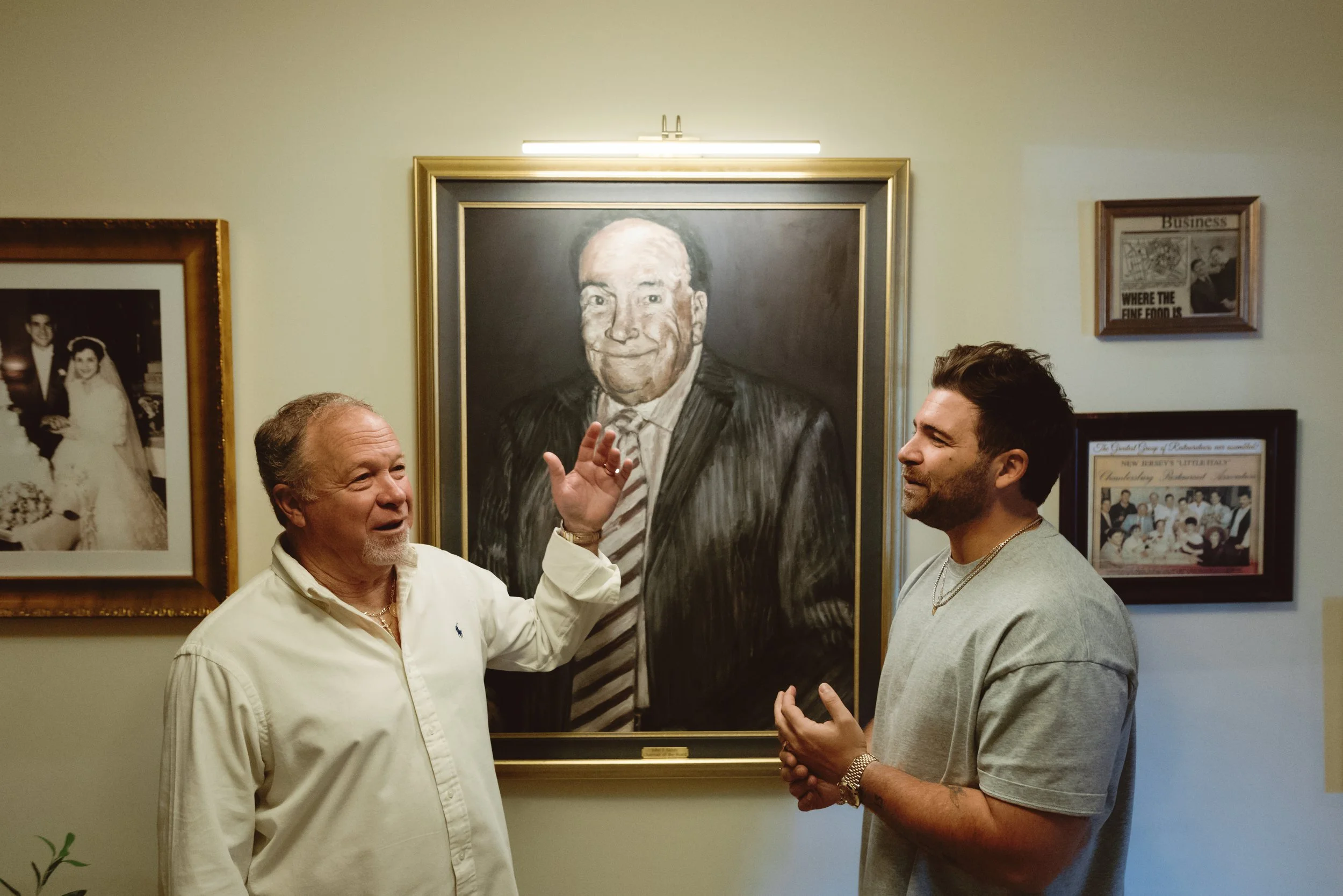 Two men talking in front of a large framed portrait of a smiling man in a suit. The man on the left is wearing a white shirt, and the man on the right is in a gray t-shirt.