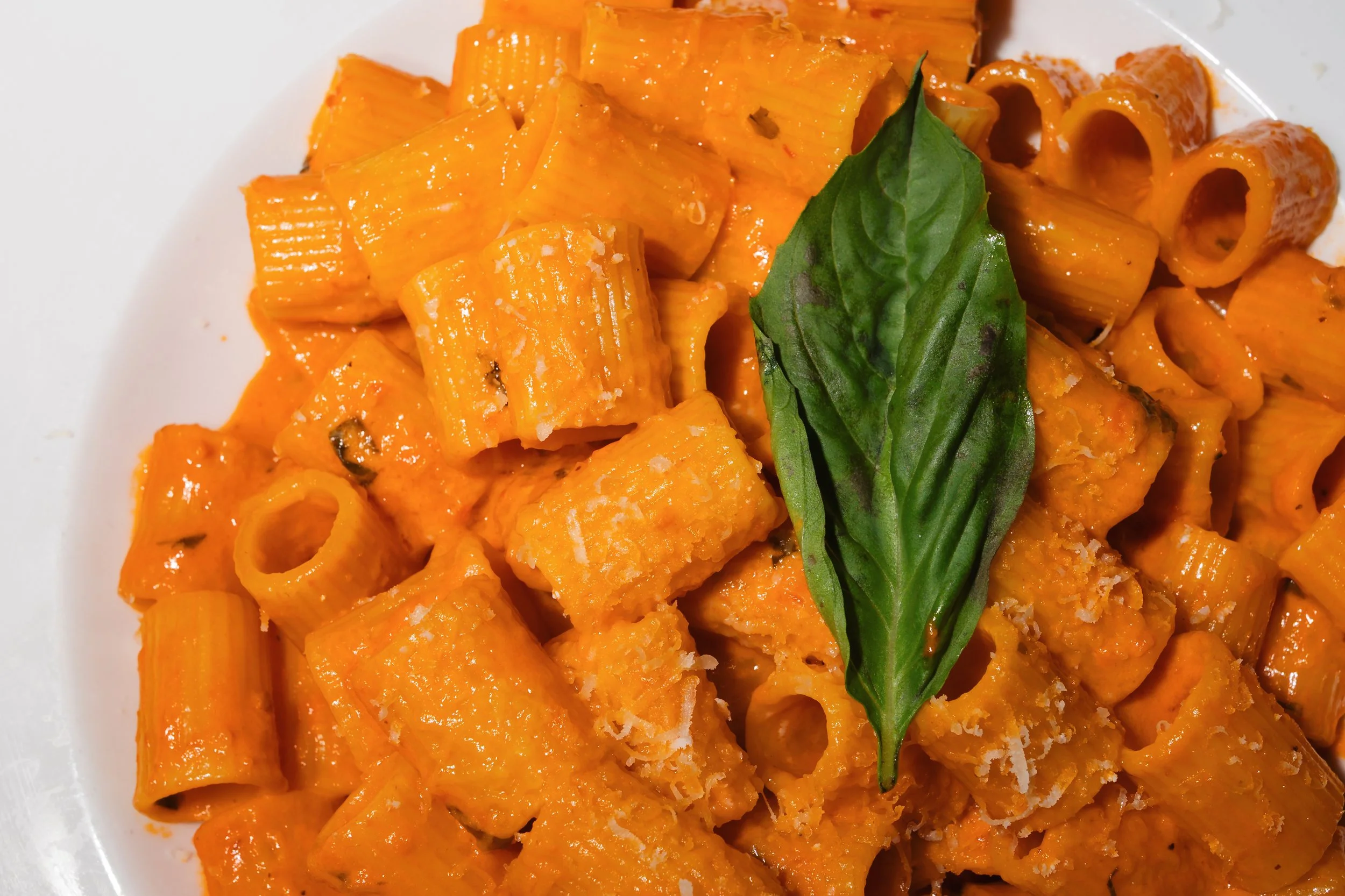Pasta with orange tomato sauce, topped with a fresh basil leaf, on a white plate.