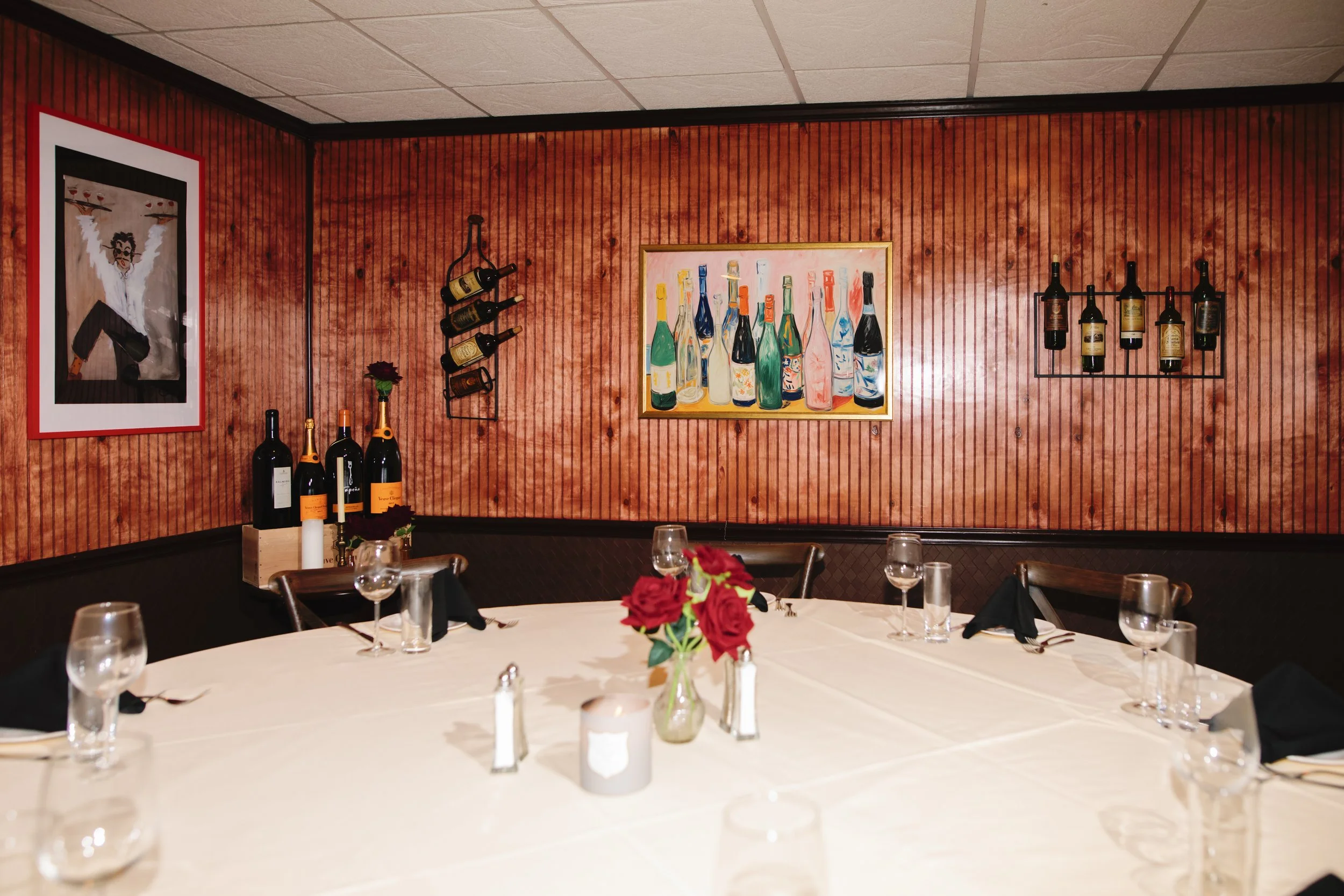 A round dining table with a white tablecloth, set with glasses, black napkins, and silverware. There is a small floral centerpiece with red roses, and a candle. The background features a red textured wall decorated with paintings of bottles and wine racks.