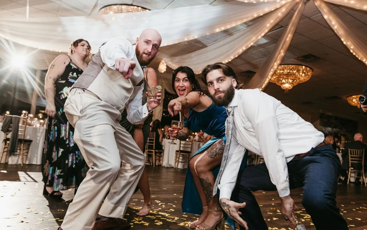 Group of people dancing and having fun at a celebration or wedding reception, with decorations and a fancy ceiling in the background.
