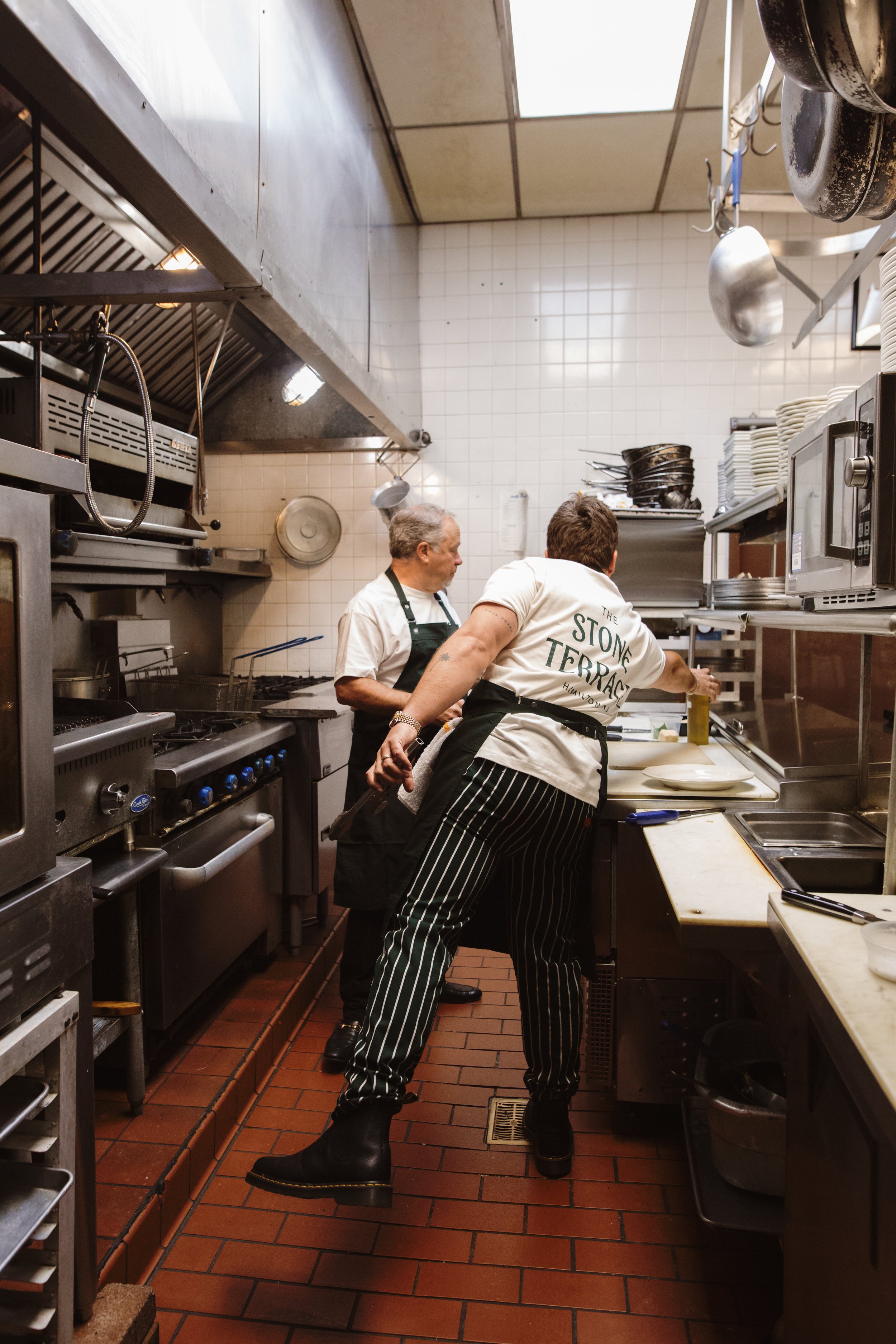 Two chefs working in a restaurant kitchen, preparing dishes at a counter with various kitchen equipment and utensils around them.