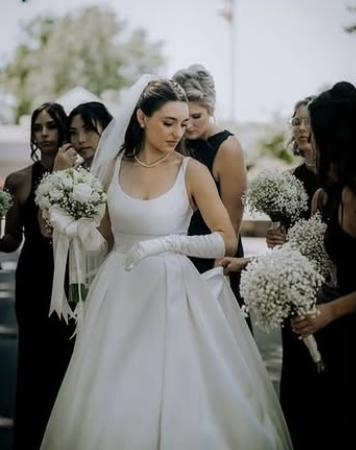 Bride in a white wedding dress and veil, surrounded by bridesmaids holding bouquets, outdoors, likely at a wedding reception.