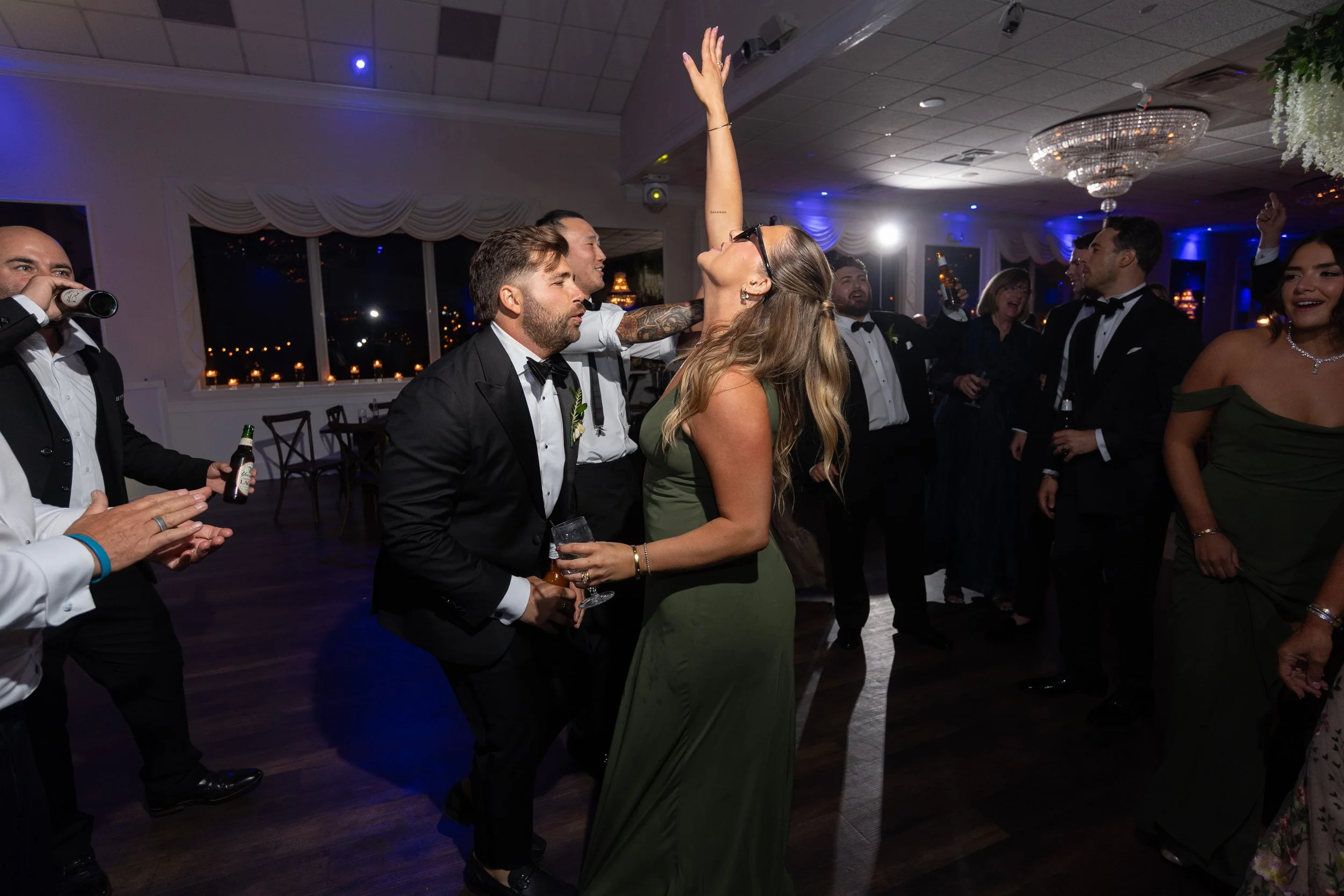People dancing and celebrating at a formal party in a decorated banquet hall with large windows and chandeliers.