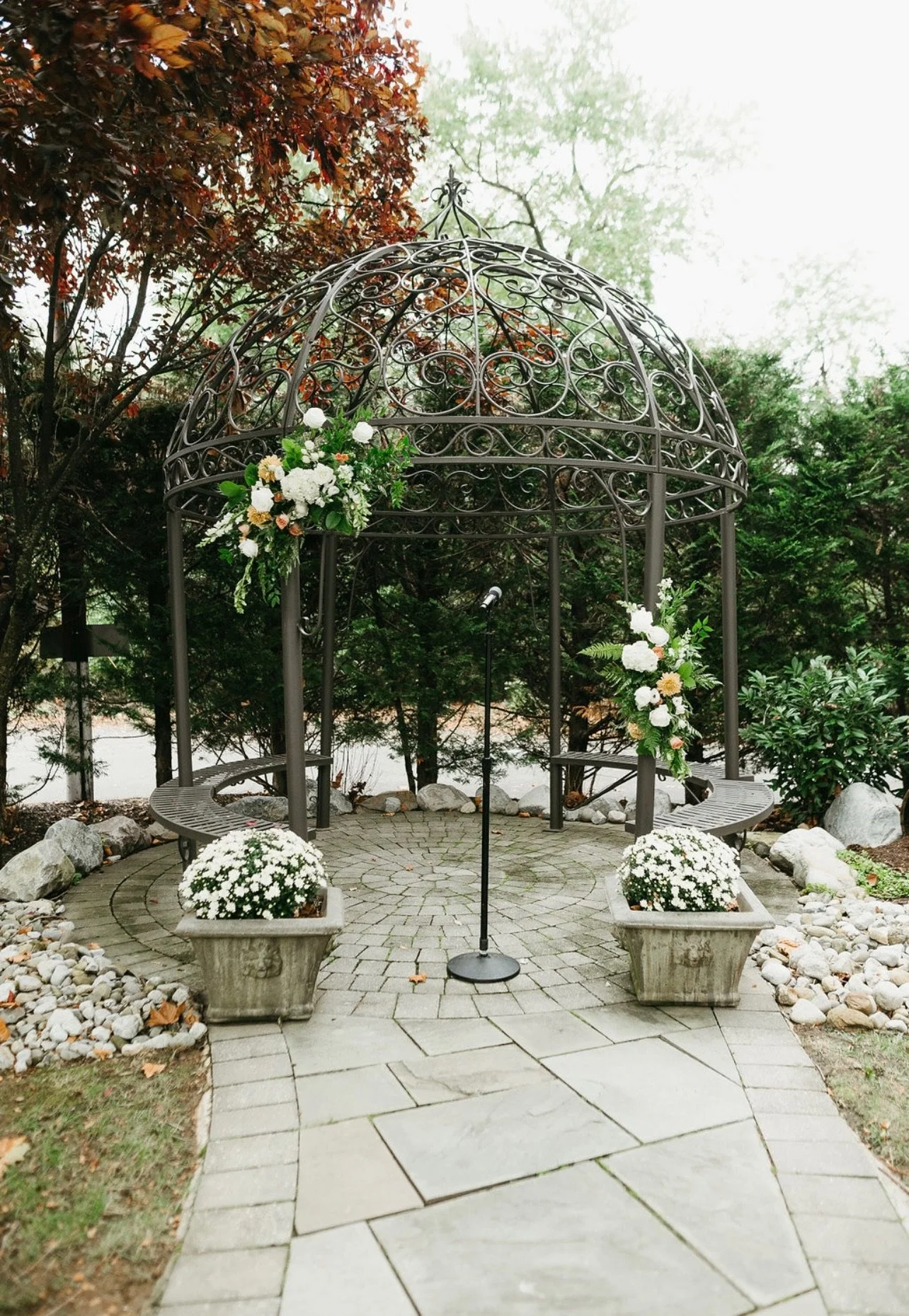 A decorative gazebo with floral arrangements, benches, and a microphone stand, set on a stone pathway amidst trees and greenery.