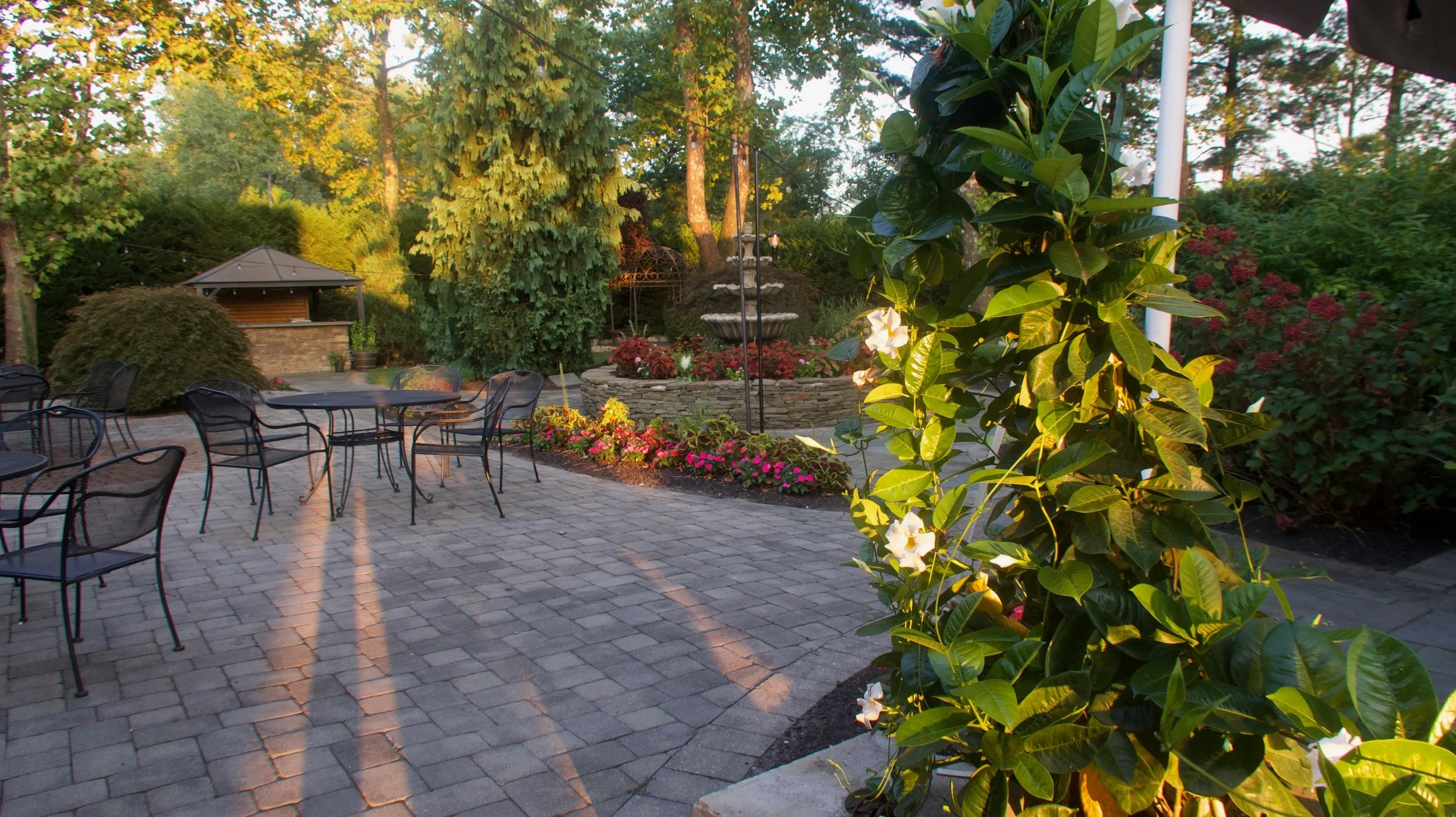 Outdoor patio with black metal chairs and a table, surrounded by colorful flowers, green trees, and a multi-tiered fountain in the background.
