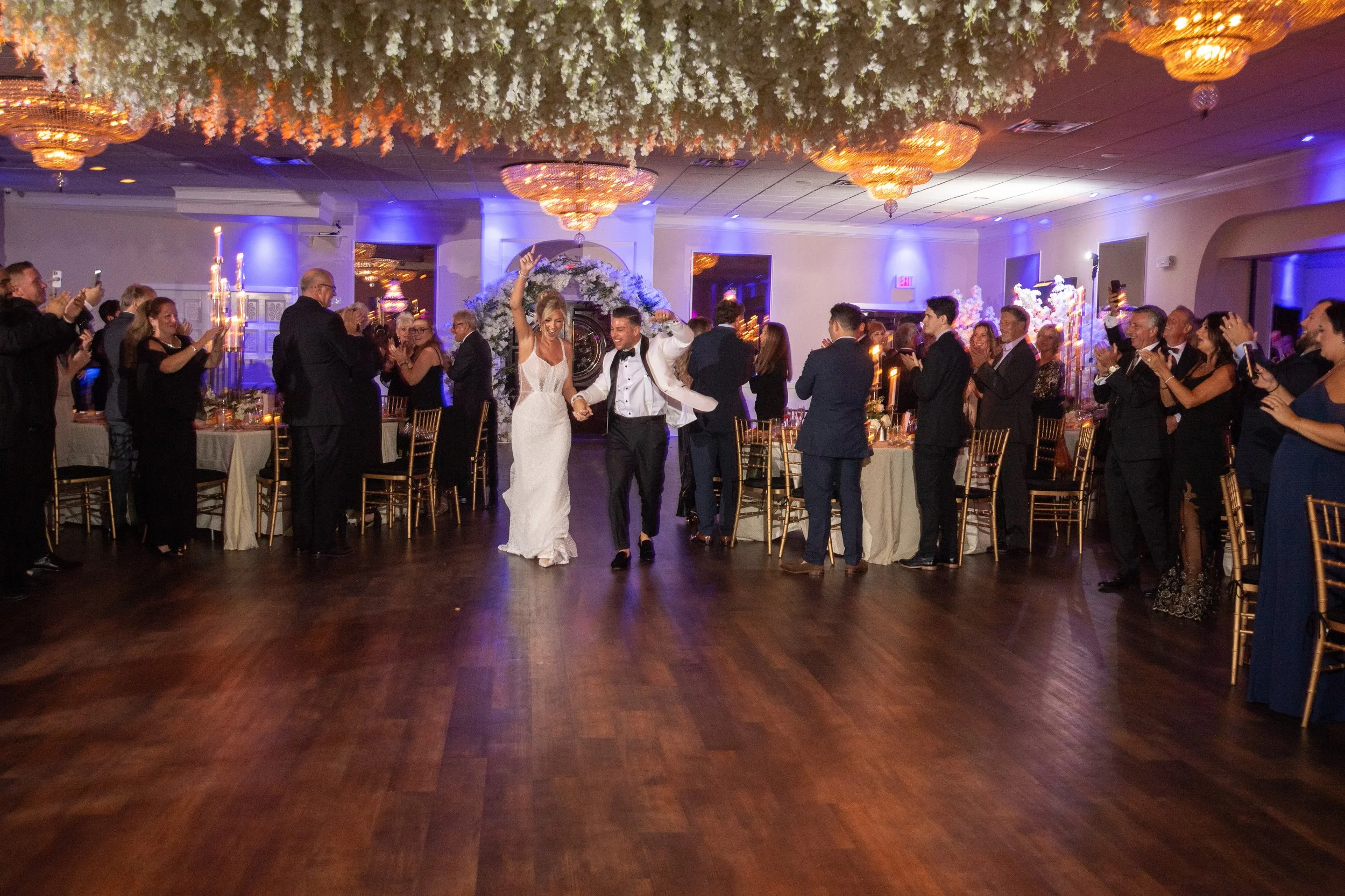 Bride and groom dancing at their wedding reception, surrounded by guests taking photos and clapping, inside a decorated banquet hall.