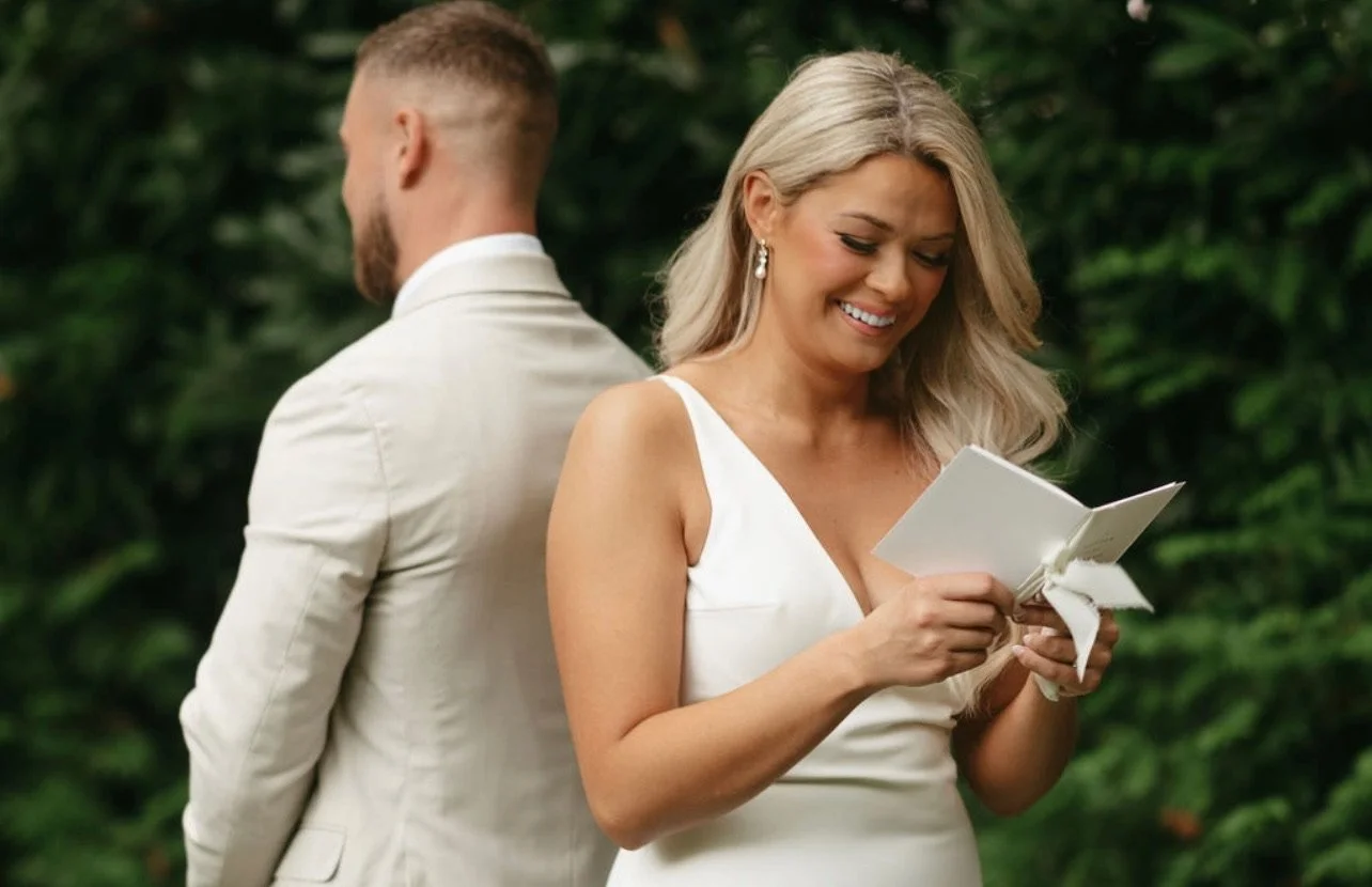A bride in a white dress smiles while reading a card, standing outdoors with a groom in a light-colored suit in the background, surrounded by green foliage.